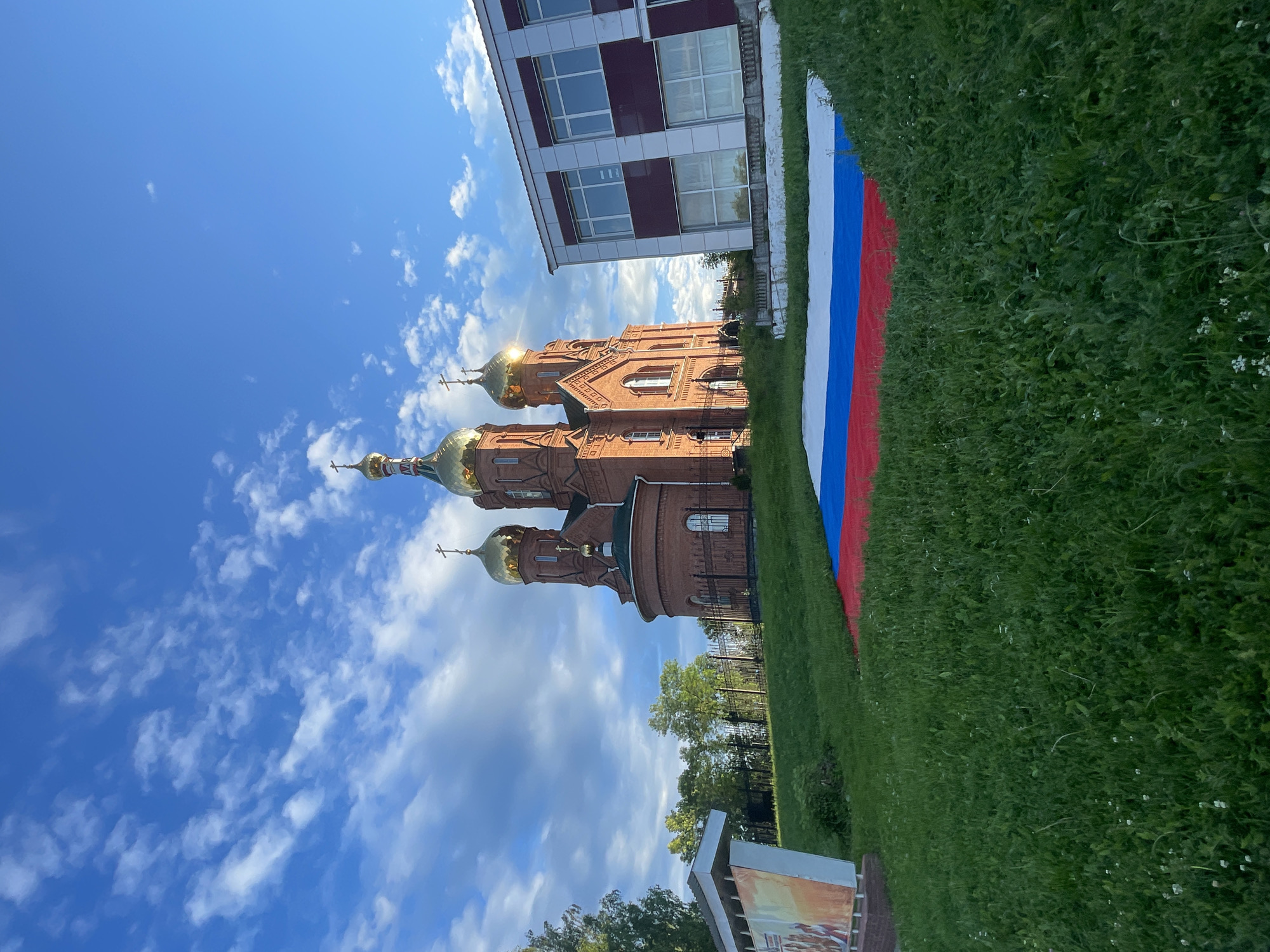 The image depicts a Russian Orthodox church with golden domes, constructed of red brick, situated on a grassy hillside. In the foreground, a large Russian flag is spread across the lawn. Adjacent to the church on the right is a modern building with a white and maroon facade and large windows. On the left side of the image, a white informational stand with colorful graphics is visible. The sky above is a clear blue with scattered white clouds. The scene is set in Krasnye Baki, Russia.
