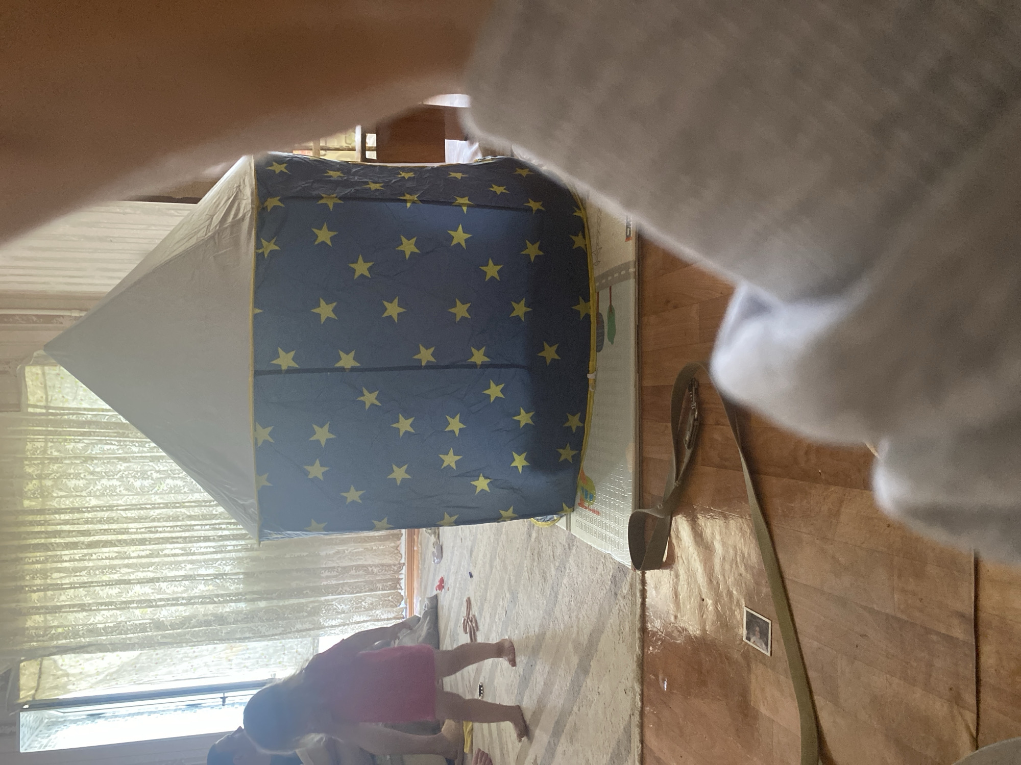 A young child, wearing a pink dress, stands on a rug in what appears to be a room. To the right of the child is a blue tent with yellow stars, partially obscured by a light-colored object in the foreground. In the background, sheer curtains hang by a window. On the wooden floor in front of the tent lies a tan strap with a buckle. A small, framed photograph is visible on the floor near the strap. The scene suggests an indoor domestic setting, likely a child's play area. There are no discernible details about the city of Shakhtinsk, Kazakhstan, in this image.