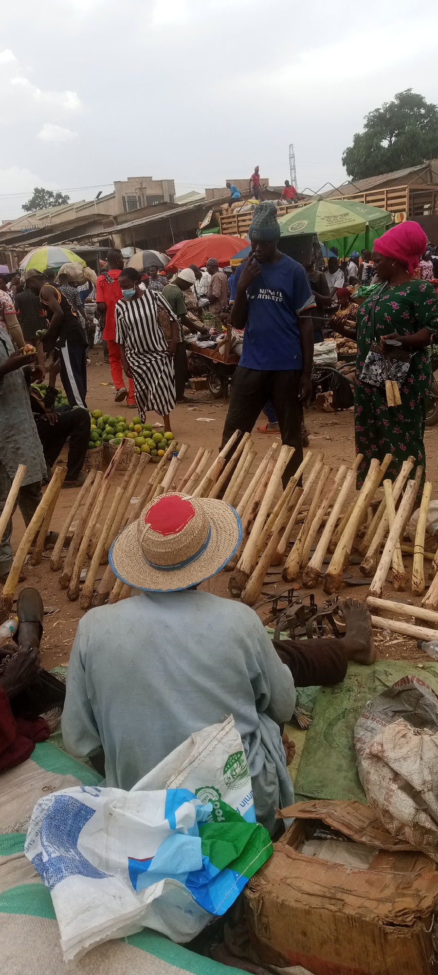 The image depicts a bustling marketplace in Gura Top, Nigeria. The foreground is dominated by a seated vendor wearing a straw hat with a red patch on top, his back to the viewer. Before him are dozens of wooden poles, arranged vertically, possibly for sale. Scattered around him are bags and a cardboard box.
In the middle ground, numerous people mill about the market. One man wears a blue t-shirt and a beanie, while a woman in a green floral dress and a pink headscarf appears to be making a purchase. Another individual wears a black and white striped dress. A pile of green fruits, possibly oranges, sits on the ground near a customer.
The background shows a variety of market stalls under red and green umbrellas. Some people are on rooftops of the structures, and others appear to be unloading materials from the back of a truck. The overall impression is one of a busy, active trading environment. The sky is overcast.