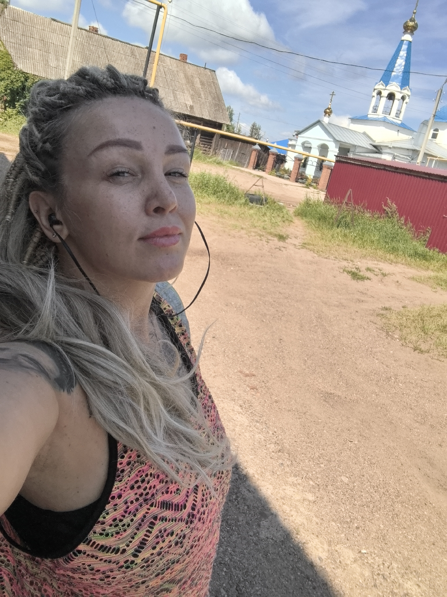 A woman with blonde dreadlocks and a pink and orange open-knit top is captured in a selfie outdoors. She is wearing black earbuds. In the background, a dirt road leads towards a blue-domed white church with a golden cross on top, situated in Noviy, Russia. To the left of the church, a rustic wooden building with a corrugated roof stands. A red corrugated metal fence borders the right side of the scene. The sky is blue with scattered white clouds. The environment appears to be rural or semi-rural.