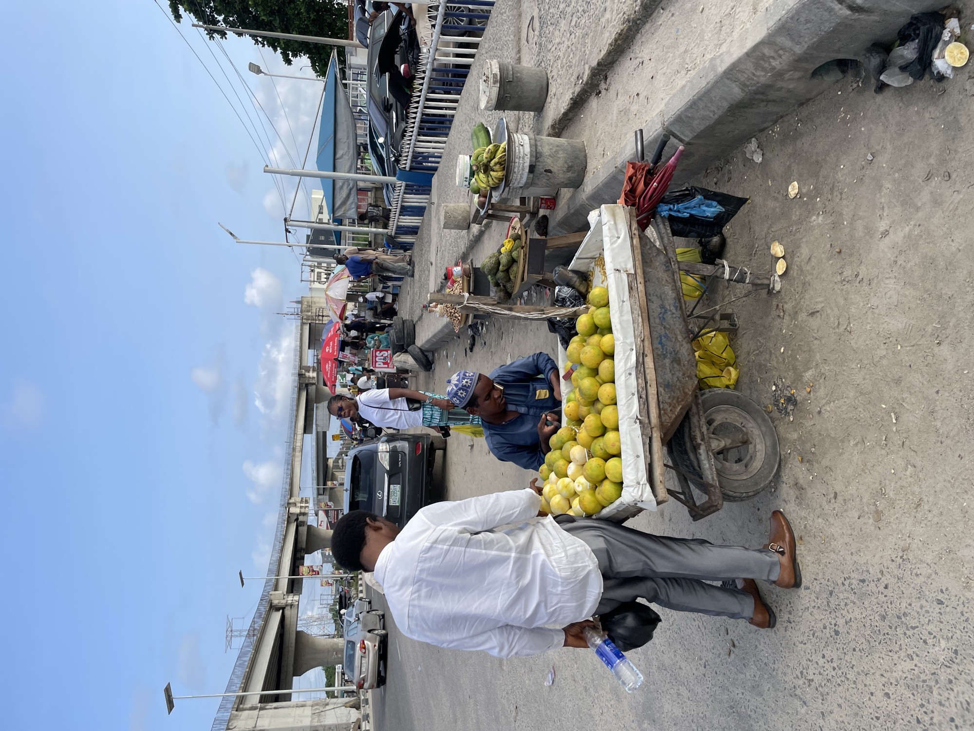 This image depicts a street scene in Lekki, Nigeria, with an overhead bridge dominating the background. In the foreground, a street vendor is selling oranges from a cart. A customer, dressed in a white shirt and grey trousers, is looking at the oranges. The vendor, wearing a blue traditional outfit and a cap, appears to be attending to the oranges. Several vehicles are visible on the road, suggesting an urban environment. The area around the vendor's cart is cluttered with other goods for sale, including bananas and other produce, as well as some debris on the ground. The sky is partly cloudy.