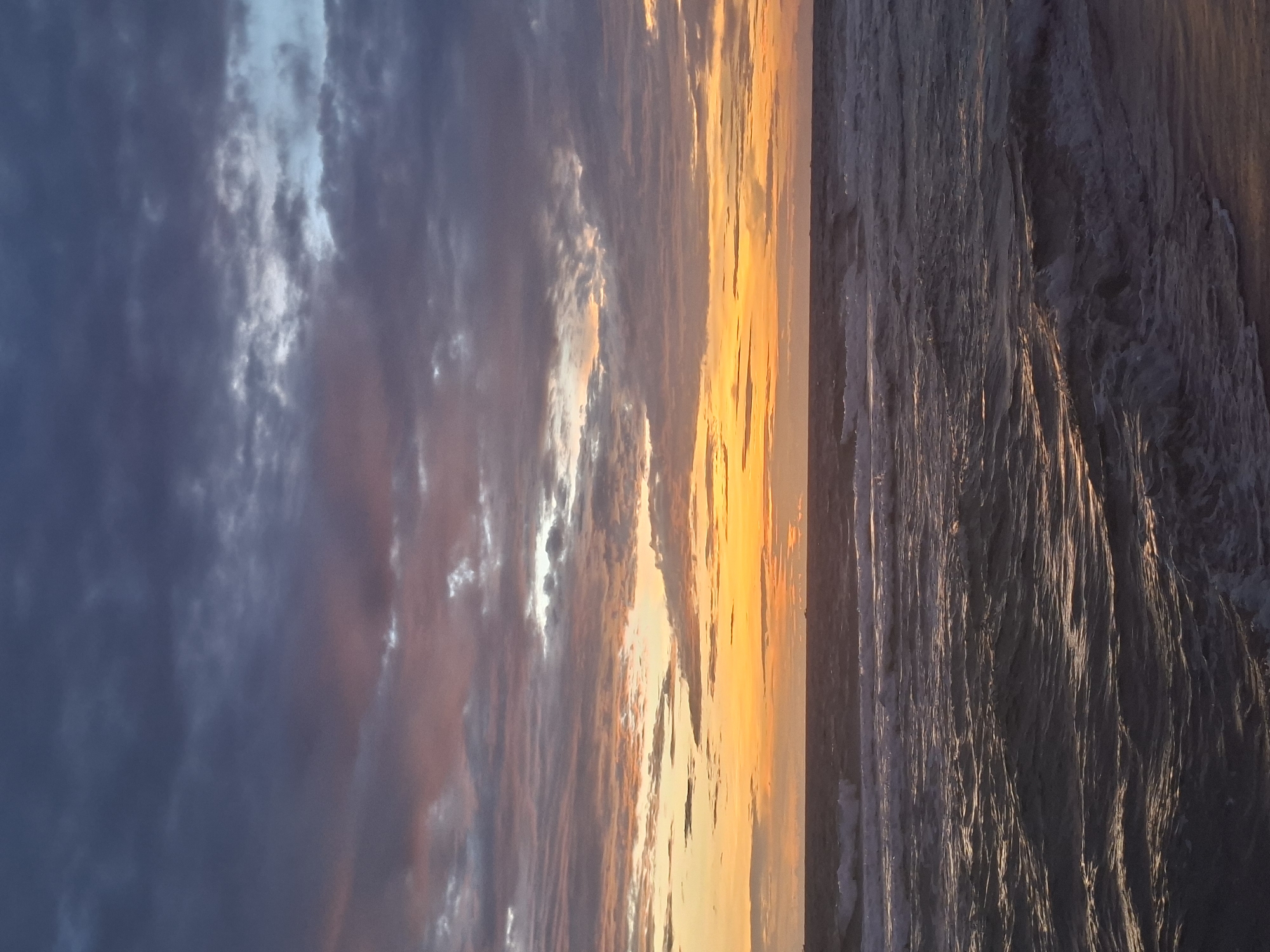 This vertical, eye-level shot captures a dramatic seascape at sunset or sunrise on Phú Quốc, Vietnam. The sky dominates the upper two-thirds of the frame, filled with a turbulent display of clouds. Dark, moody grays and deep purples transition to softer pinks and vibrant oranges near the horizon, indicating the fading or emerging sunlight. Glimmers of lighter sky peek through breaks in the cloud cover, adding texture and depth.
Below the sky, the ocean stretches across the bottom third of the image. The water is choppy, with whitecaps and foam suggesting moderate wave action. The surface reflects the colors of the sky, with streaks of orange and gold shimmering on the darker, textured water. The horizon line is a subtle band separating the sky and sea, with distant, faint shapes that could be boats or landmasses. The lower edge of the frame shows a sliver of dark sand, hinting at a beach. The overall atmosphere is one of natural beauty and powerful, dynamic weather.
