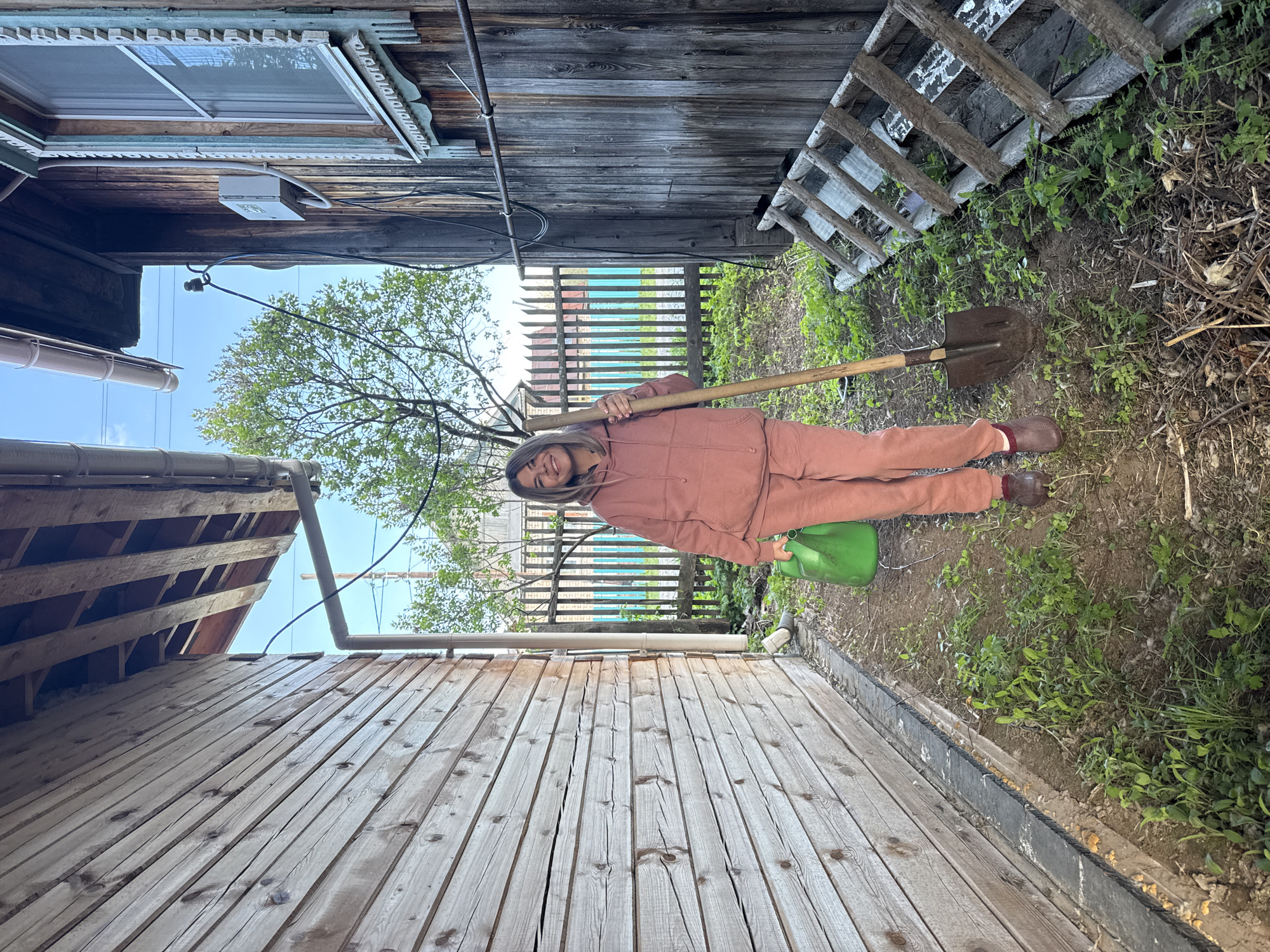 A woman stands in an outdoor space between two wooden buildings in Chaikovsky, Russia. She wears a matching pink hoodie and pants with brown shoes. She holds a shovel over her shoulder and has a green watering can beside her feet. A wooden ladder leans against one building. A blue and white fence is visible in the background. The sky is clear.