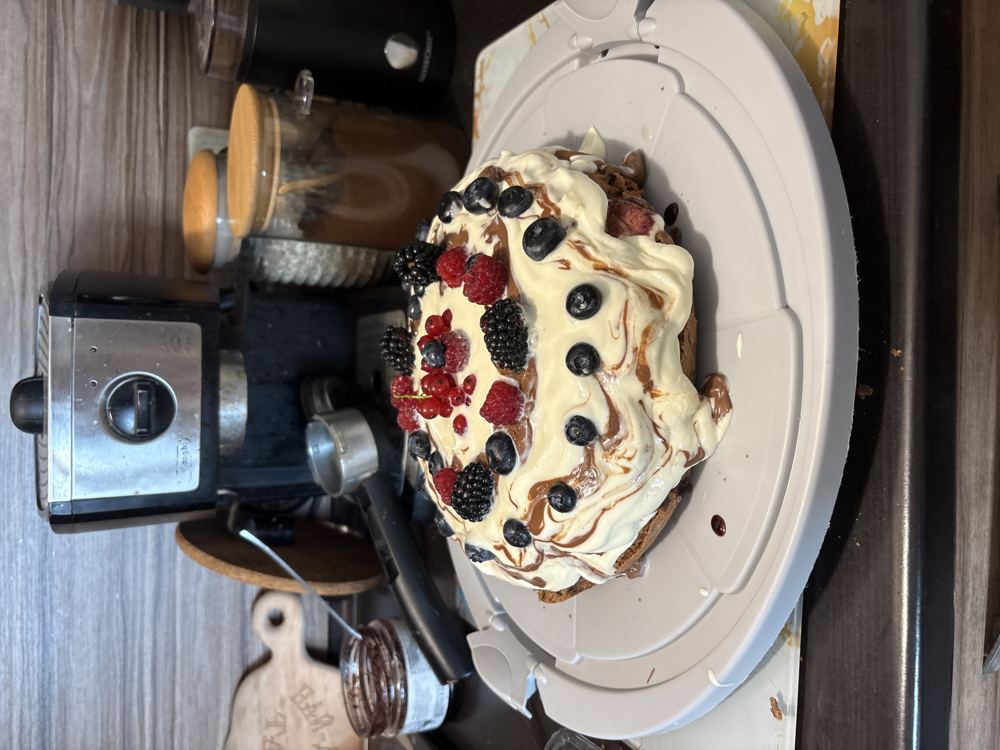 A close-up, eye-level shot shows a frosted cake topped with berries, including raspberries, blueberries, blackberries, and red currants, all arranged on a grey cake stand. The cake is drizzled with a chocolate-colored sauce. In the background, a metallic espresso machine is visible, along with two glass jars containing what appears to be coffee beans and cocoa powder. To the left, a small glass jar of jam and a wooden cutting board are also present. The scene is set in a kitchen environment with a wooden-paneled wall.