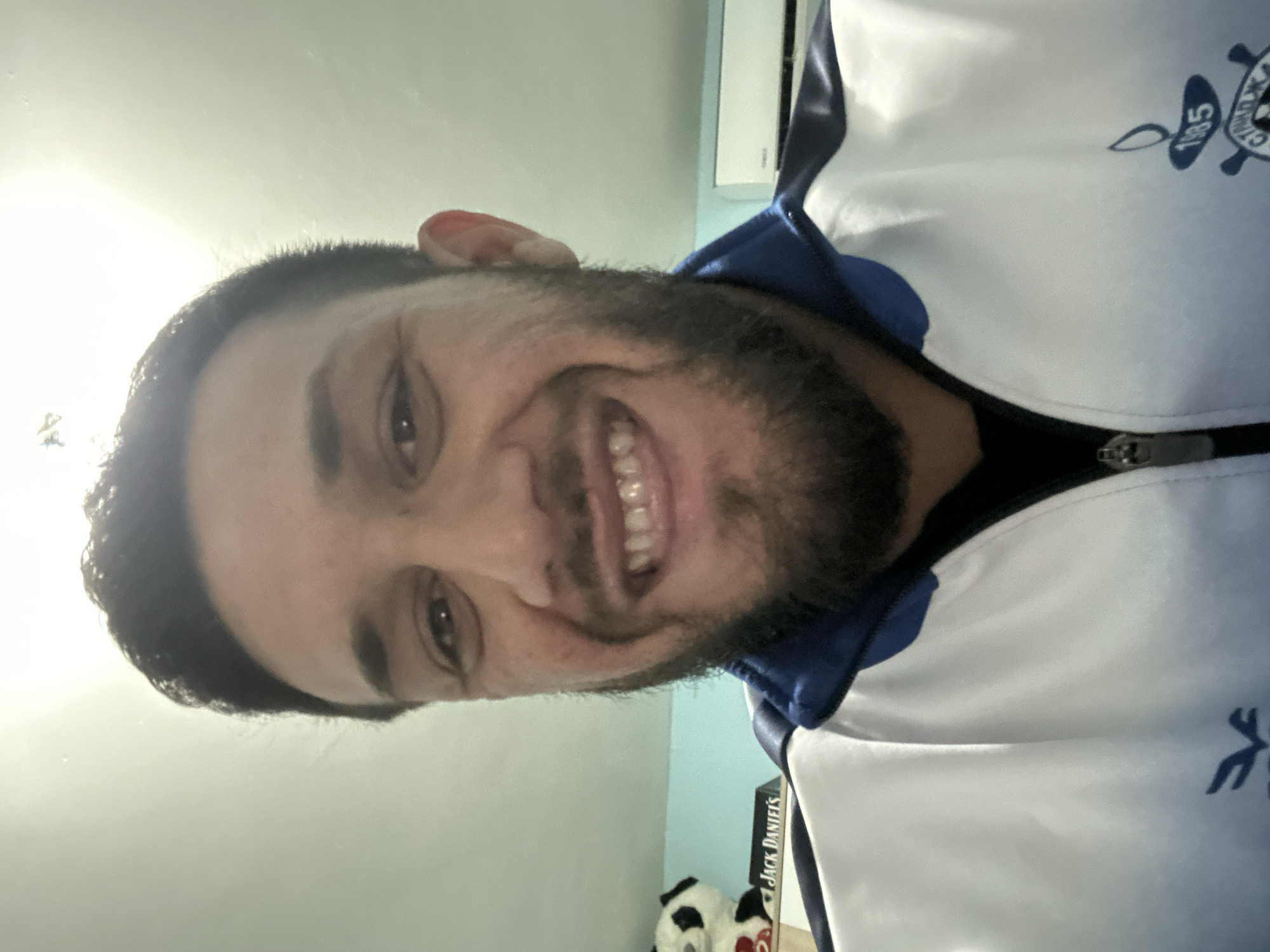 A man with dark hair and a beard is smiling at the camera. He is wearing a white and blue jacket with a logo on the upper right side and a zipper in the middle. Behind him is a light blue wall. A white air conditioner is visible in the upper right corner of the image. A bottle of Jack Daniel's whiskey and a stuffed panda are also visible in the lower portion of the image. The location, Tsarevo, Bulgaria, cannot be determined from the image.