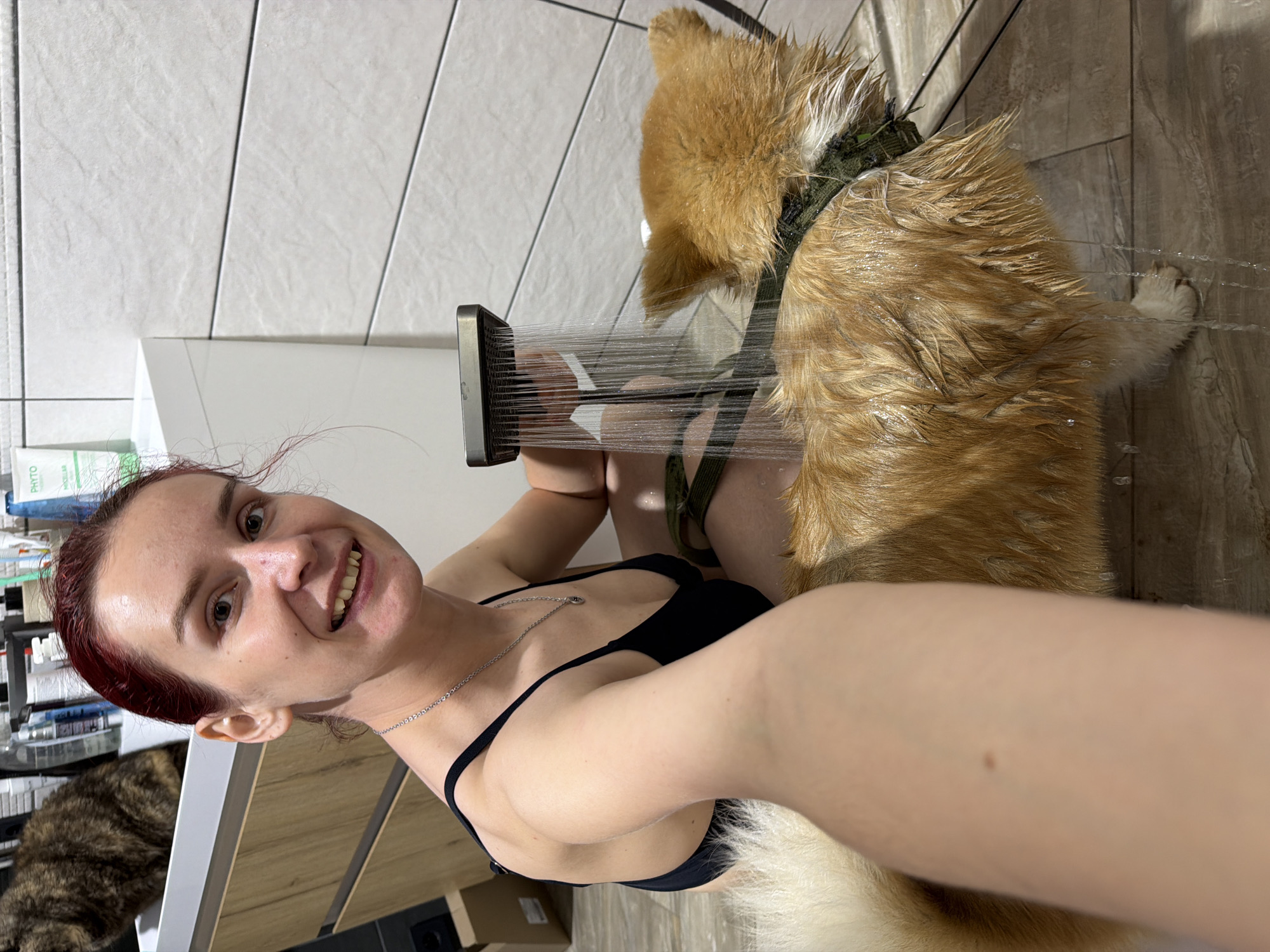 A woman with red hair, wearing a black top and a silver necklace, is holding a showerhead and smiling at the camera while giving a dog a bath. The dog, which is brown and white, is wearing a green harness. Water is spraying from the showerhead onto the dog. In the background, there are white and beige tiles, shelves with toiletries, and a cat. The location is Chaikovsky, Russia.