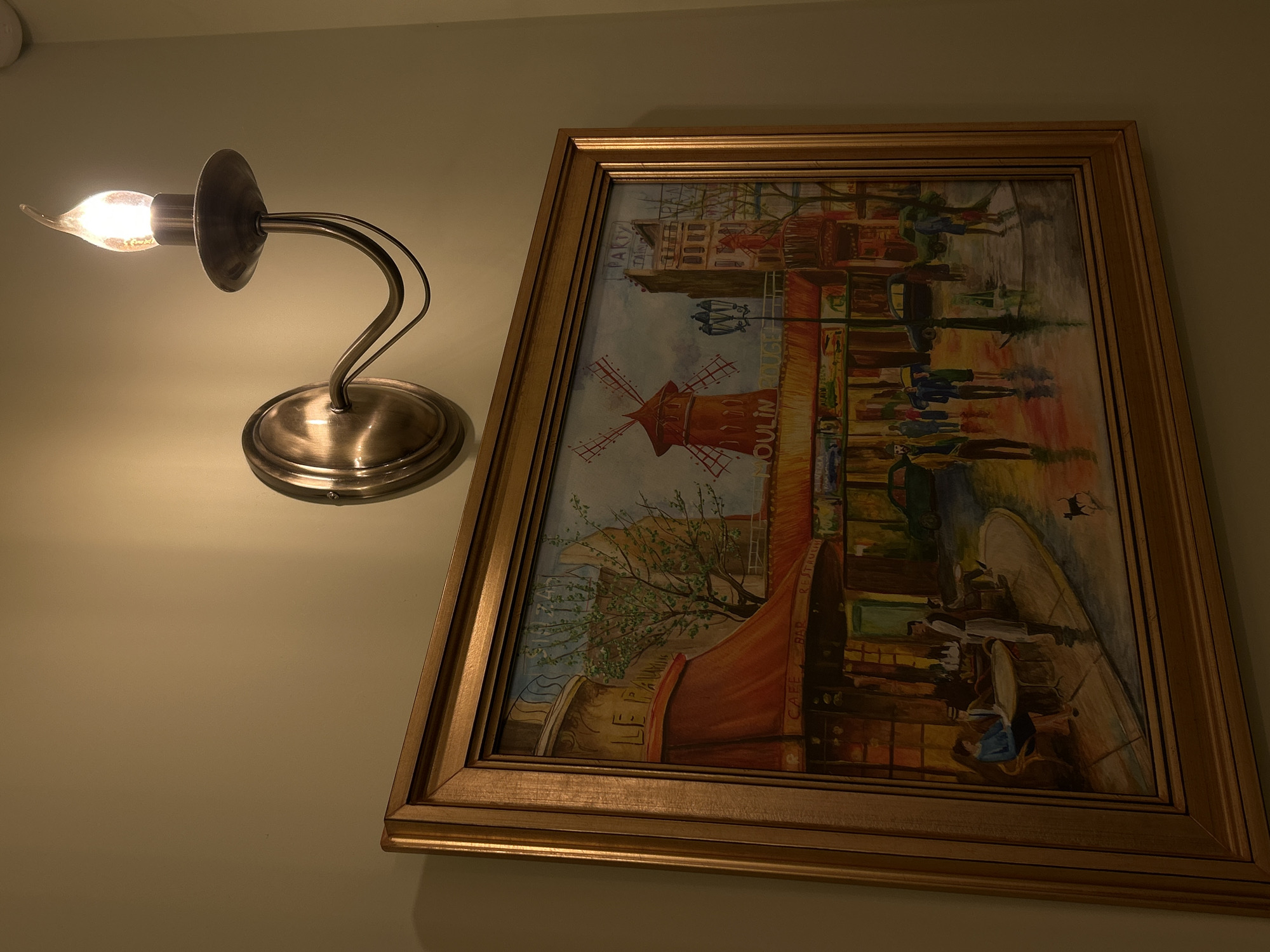 A framed painting of a Parisian street scene hangs on a light-colored wall next to a sconce. The painting depicts a cafe scene with figures seated at tables and pedestrians walking along the street. A windmill is in the background. The sconce has a silver-colored base, a curved arm, and a flame-shaped bulb. The light from the bulb illuminates the painting and wall.