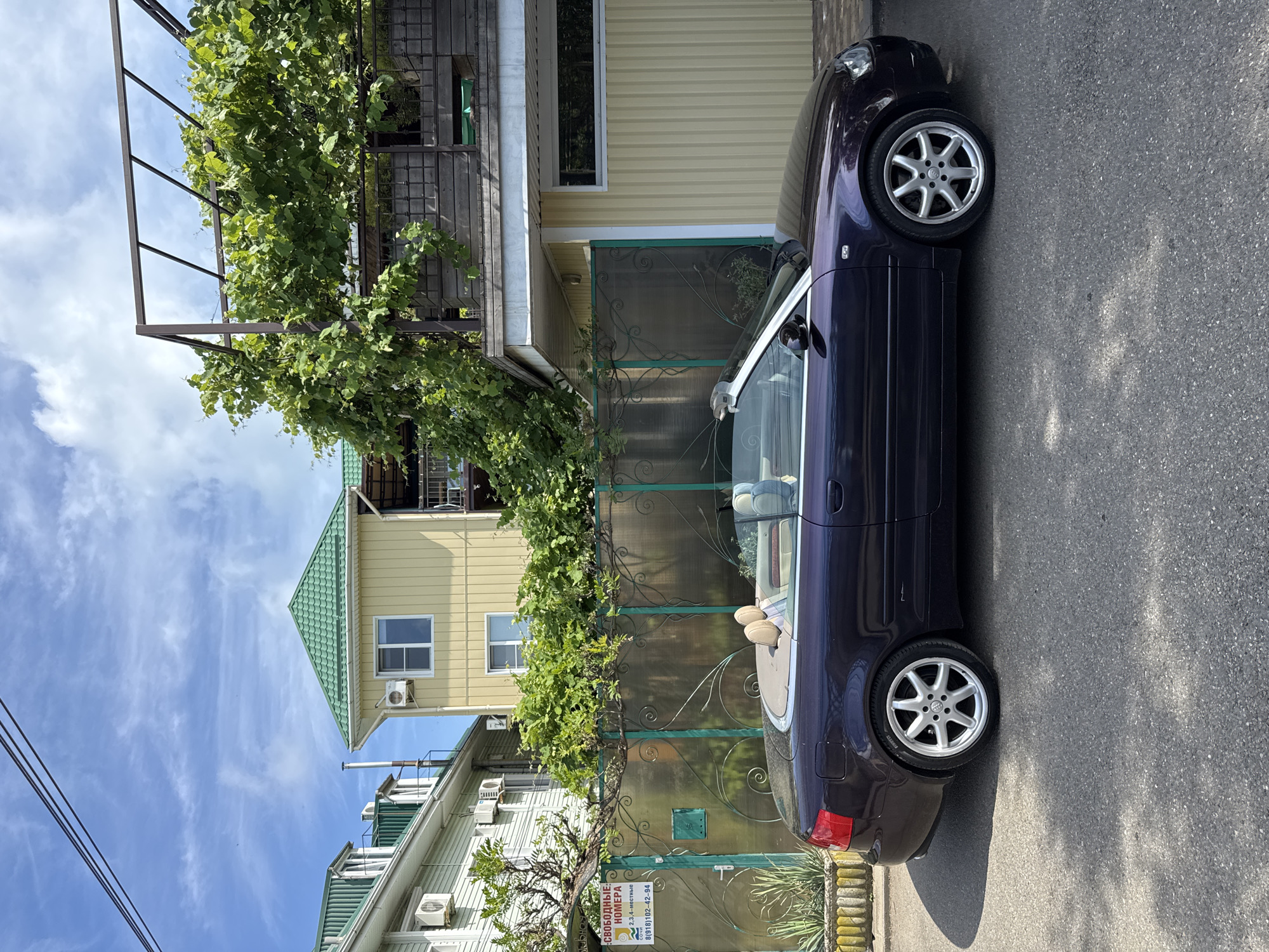 A dark purple convertible Audi A4 is parked on a paved road in front of a metal fence with ornate scrollwork, behind which are two residential buildings. The building on the left is light green with white trim and air conditioning units visible on the exterior. The building on the right is yellow with a green roof and a large section of overgrown green foliage on a trellis structure. A sign with Russian text indicating "FREE ROOMS" is attached to the fence. The sky is blue with scattered white clouds, and power lines are visible in the upper left quadrant of the image. The scene appears to be a residential area in Sochi, Russia, likely during daylight hours.