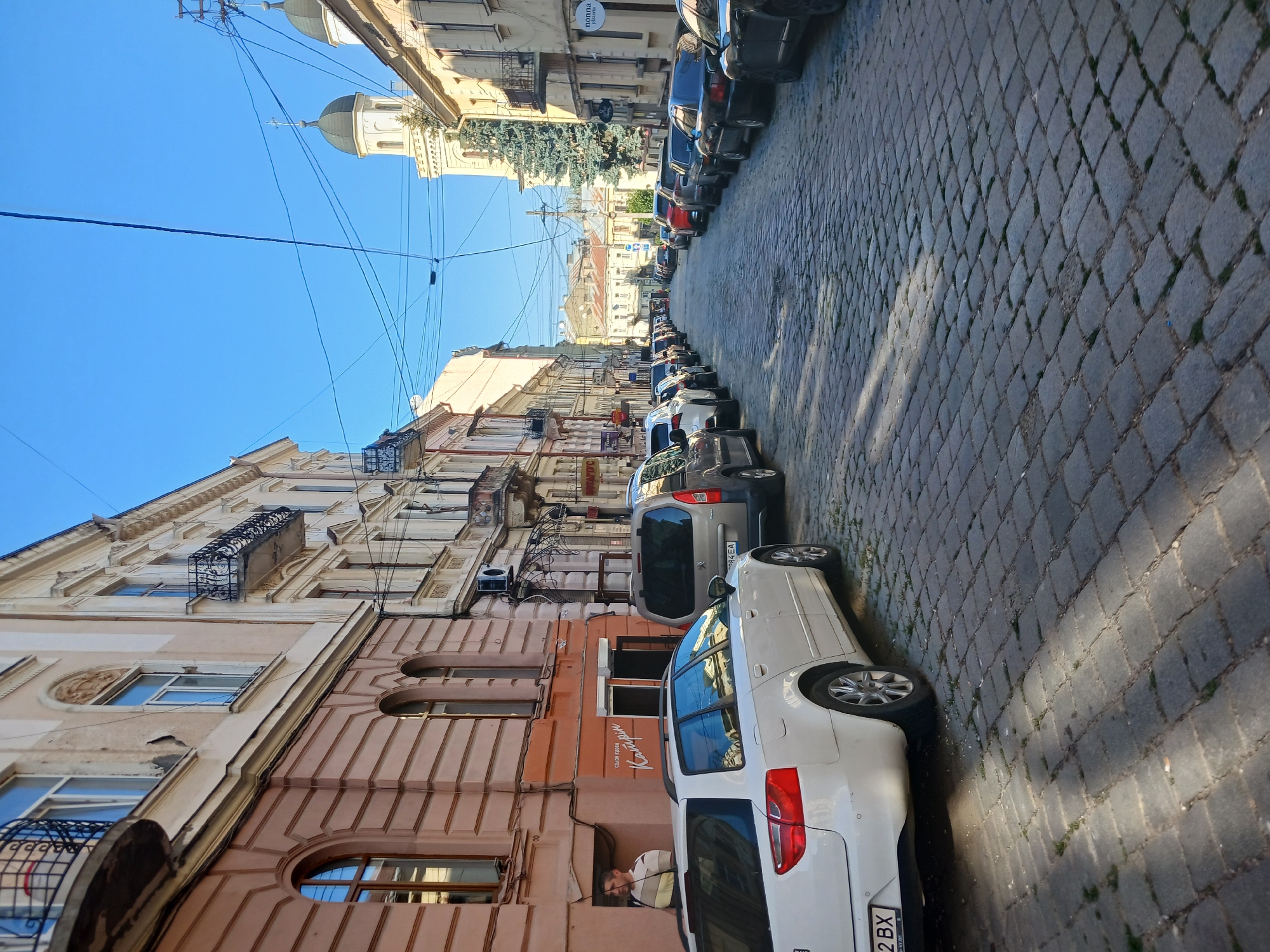 The image depicts a street in Chernivtsi, Ukraine, characterized by its cobblestone pavement and historic architecture. On the left side of the street, a man wearing a striped shirt stands near the entrance of a building with a sign that reads "Салон краси Катрін" (Beauty Salon Katrin). Several cars are parked along the street, including a white station wagon in the foreground. The buildings lining the street are multi-story structures with ornate balconies and detailed facades, indicative of old European architecture. In the background, two church domes are visible, suggesting the presence of religious institutions within the city. The sky is clear and blue, and sunlight casts shadows on the street, indicating it is daytime. The scene appears to be a typical urban streetscape in the historic center of Chernivtsi.