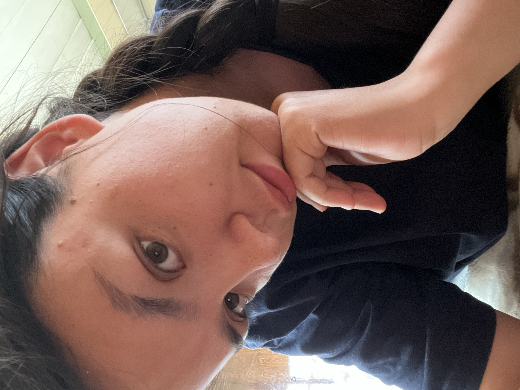 A single individual, appearing to be a young adult female, is depicted in a close-up, upward-angled indoor portrait. The subject's head is tilted to her right, and her right hand is positioned under her chin, supporting her jawline. She looks directly at the camera with dark eyes. Her dark hair is partially visible, with a braid resting over her right shoulder. Her skin displays natural textures, including visible pores and minor blemishes on her forehead and cheek. She is wearing a plain, dark-colored (likely navy blue or black) short-sleeved garment. In the background, a bright, diffused light source is visible to the left, possibly from a window or door. To the upper right, a ceiling structure with light-colored rectangular panels and darker borders or accents is partially visible. Below the subject's right arm, a light-colored, soft-textured surface, possibly a blanket or rug, is present. The specific location in the city of Oktyabrsky, Russia, cannot be determined from the visual information present in the image.