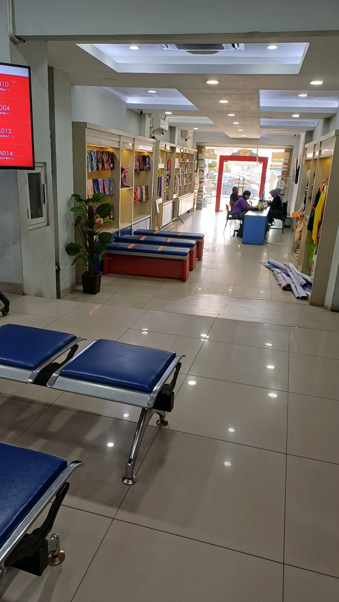 This image shows the interior of a store located in Cibinong, Indonesia. In the foreground, two rows of blue benches with metal frames are visible, set against a backdrop of beige tiled flooring. The benches appear to be part of a waiting area.
The middle ground of the image features several shelves displaying various textile products, possibly fabrics or garments, in a retail setting. A potted plant stands next to the shelves. Further back, a counter area is set up with two individuals, one appearing to be a staff member and the other a customer, engaged in an interaction. Behind them, a red doorway is visible, with a glimpse of the outdoors, suggesting the store's entrance.
The environment is well-lit with overhead lights, and the overall impression is that of a commercial establishment with a focus on textile sales. The architecture of the ceiling incorporates recessed lighting and air conditioning units. In the background, to the left, a large red digital display screen is mounted on the wall, showing text and graphics, possibly indicating product information or promotions. Rolls of material are stacked on the floor on the right side of the image.