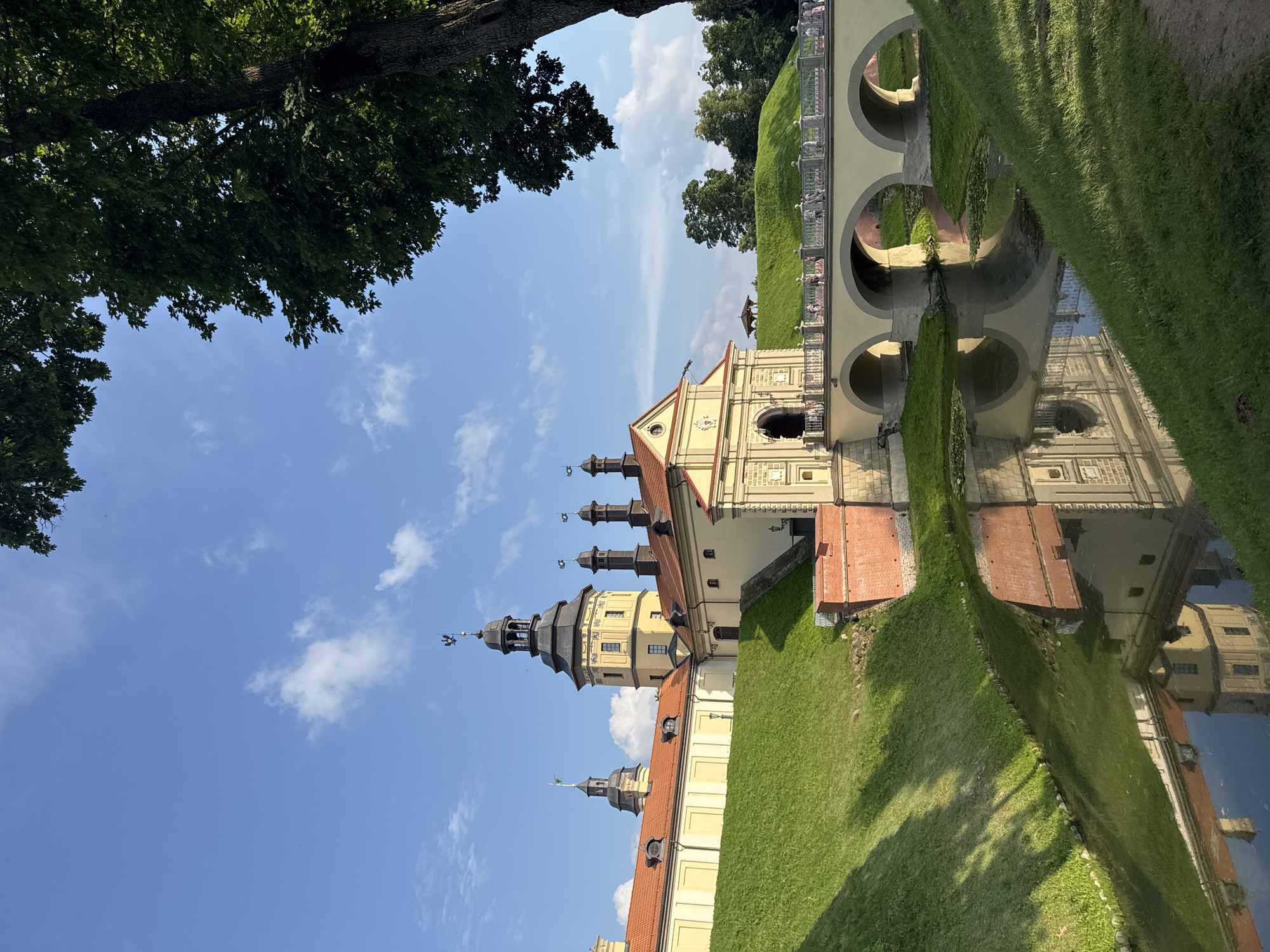 The image shows the Nesvizh Castle complex in Nesvizh, Belarus, on a sunny day with a blue sky and scattered clouds. The castle, with its distinctive yellow walls and red tile roofs, is situated on a grassy mound. A moat with still water runs in front of the castle, reflecting the architecture above. A multi-arched bridge, lined with white railings and people, spans the moat, leading to the castle entrance. Lush green grass covers the slopes of the mound and surrounds the moat. A large tree on the right side of the frame partially obstructs the view of the sky and castle. The scene is one of historical grandeur and natural beauty.