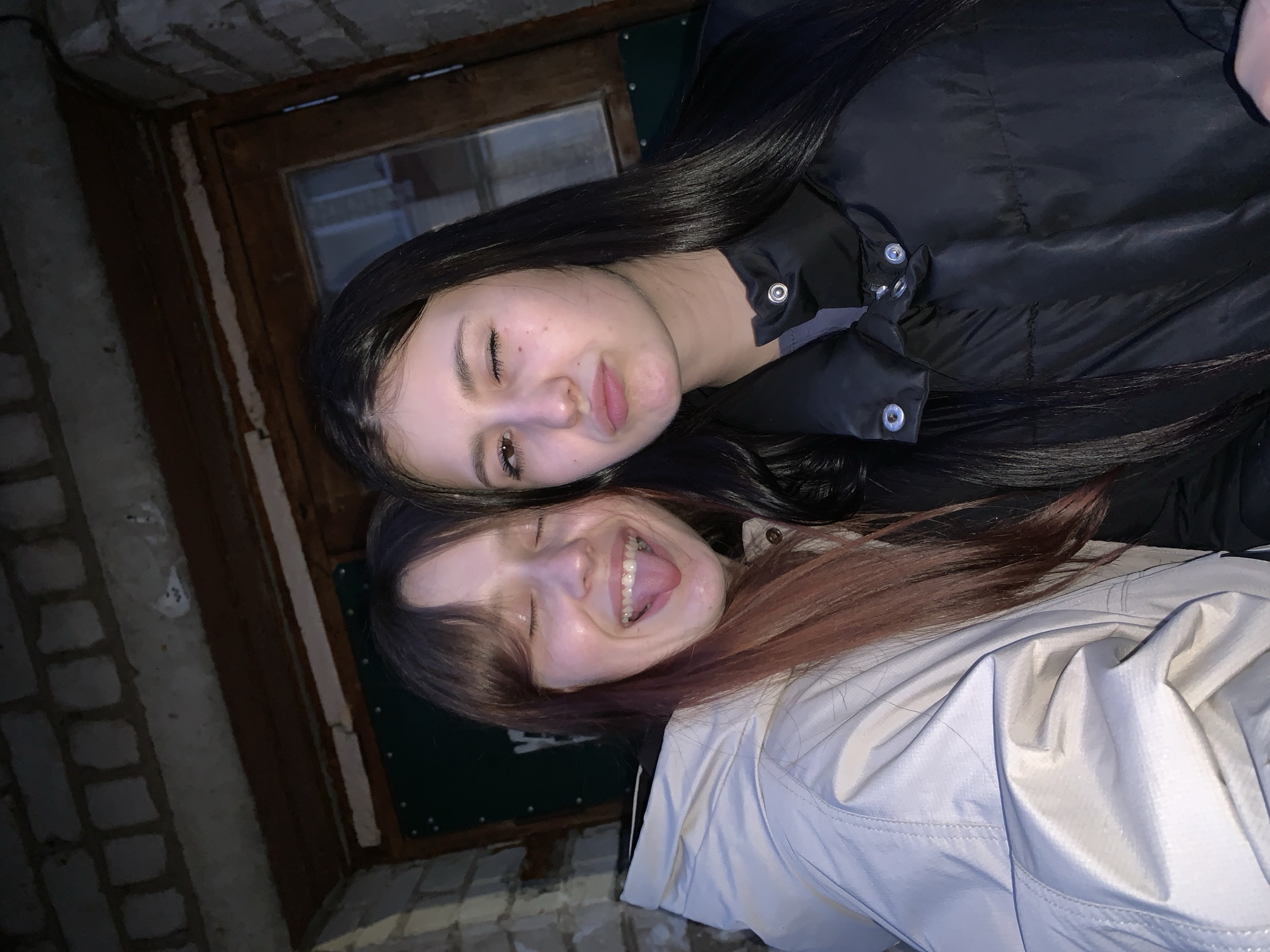 A low-light, candid portrait captures two young women posing playfully, likely outdoors, against a rustic architectural background. The image is rotated, with the subjects appearing diagonally across the frame.
The woman at the top has long, straight dark hair and is wearing a black puffer jacket. She has a subtle smile and her eyes are partially closed, as if winking or squinting. Her skin appears fair, and she has visible freckles or moles on her cheek.
Below her, and slightly in front, is another woman with reddish-brown hair, styled with bangs, and wearing a light-colored, possibly white or light grey, jacket with a textured surface. She is smiling broadly with her eyes closed, playfully sticking out her tongue. Both women appear happy and engaged in the moment.
The background consists of dark brickwork on the left and top, and a wooden door frame with a window pane in the center. A dark green panel is visible to the right of the door frame. The lighting suggests it is nighttime or a very dark environment, with a bright flash illuminating the subjects. The puffer jackets worn by both women suggest cool or cold weather. There are no visible texts or specific location cues beyond the architectural elements. The setting is likely an outdoor area of a building in Фоки, Russia.
