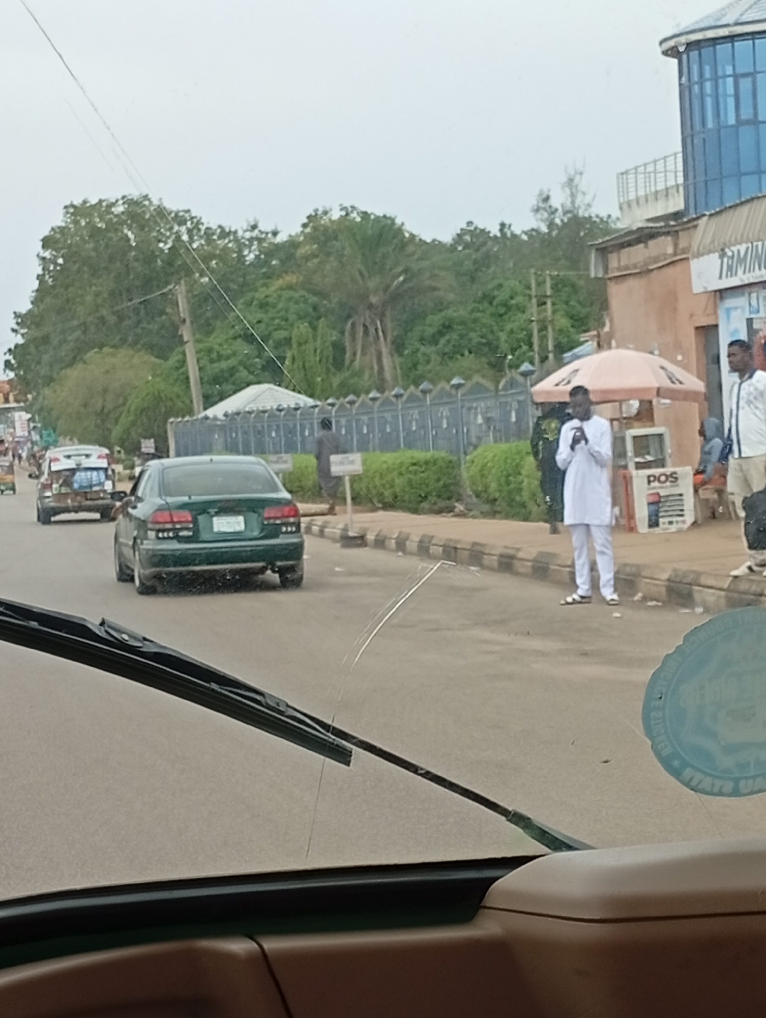 The image captures a street scene in Jos, Nigeria. In the foreground, the windshield and dashboard of a vehicle are visible, suggesting the photo was taken from inside another car. The road itself is paved, with a curb and sidewalk on the right. Several vehicles are present on the road. A dark green sedan is prominently displayed in the middle ground, its rear facing the camera. Further down the road, a lighter colored car with luggage strapped to its roof is also visible.
On the sidewalk, several individuals are present. One man, dressed in white traditional attire, is looking at his phone. Nearby, another man in a white shirt and dark pants stands, while a third person, partially obscured, is seated. A stall with an umbrella and what appears to be a point-of-sale (POS) machine is set up on the sidewalk. In the background, there are buildings, including one with a distinctive blue, rounded glass facade, and lush green trees and vegetation. A fence runs along the side of the road, bordering a grassy area. The sky is overcast, contributing to a diffused light in the scene. The overall impression is of a busy, ordinary day on a street in Jos.
