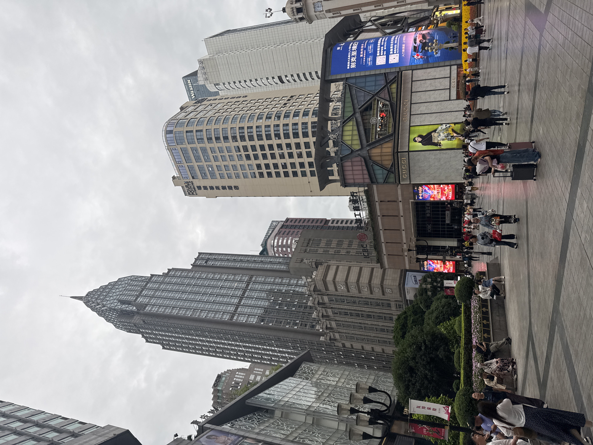 This image depicts a street scene in Yuzhong District, China, characterized by towering buildings, commercial establishments, and pedestrian activity. Several skyscrapers loom in the background, including one with a spire. Notable establishments present are Westin and Gucci, indicated by signage. Electronic advertising boards display information, including one promoting SUVs. A number of people can be seen walking and sitting on benches among planters, suggesting a public space. Suitcases indicate people might be tourists.