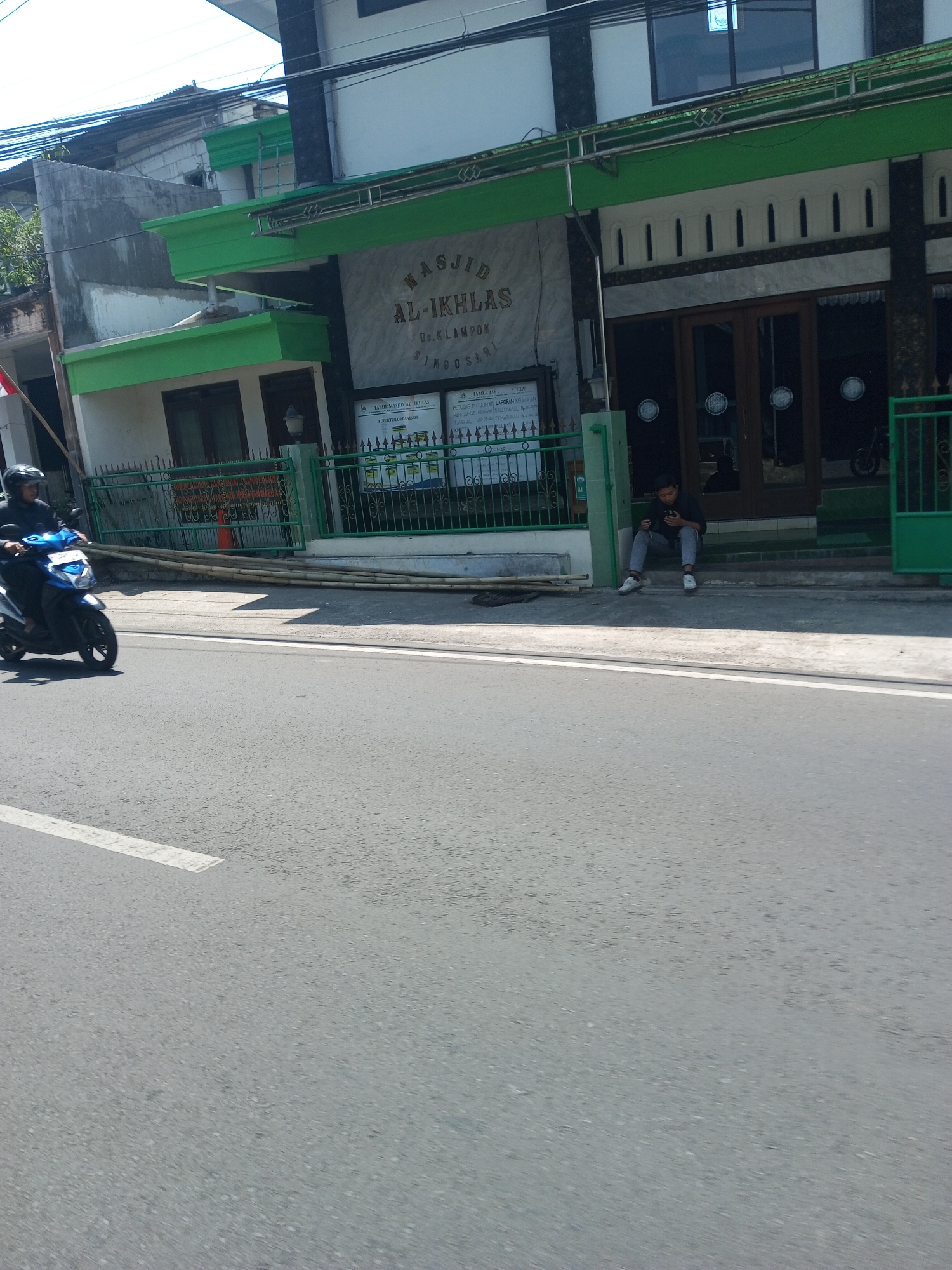 The image shows the exterior of the Al-Ikhlas Mosque in Klampok, Indonesia. In the foreground, a person is riding a blue and white scooter on a paved road. To the right, a young person is sitting on the steps of the mosque, looking at their phone. The mosque has a green trim and a sign that reads "MASJID AL-IKHLAS". Several informational boards are displayed on the wall of the mosque, and a green fence encloses the property. The sky is clear and the sun is shining.