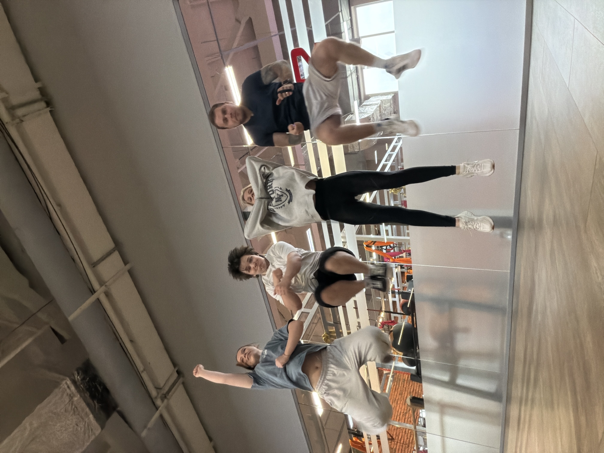 This dynamic image, taken from a low angle, features five individuals in an indoor setting, creating the illusion that they are suspended or jumping in mid-air. The scene is likely a gym or studio in Chaikovsky, Russia.
The five individuals, consisting of three men and two women, are positioned vertically across the frame. At the top, a man in a dark t-shirt and light shorts appears to float with bent knees. Below him, a woman in a light grey hoodie and dark leggings is suspended, looking towards the camera. Further down, a man in a white t-shirt and dark shorts is mid-pose. Towards the bottom, a woman in a light blue t-shirt and grey sweatpants strikes another dynamic pose, while a man is partially visible at the far bottom left in light athletic wear. All subjects are dressed in casual or athletic attire.
The background primarily consists of a high ceiling with exposed grey ducts and pipes running across the left side, characteristic of a large commercial or industrial space. A significant portion of the right side of the image is dominated by large, reflective panels, likely mirrors, which create complex reflections of the interior. These reflections include glimpses of what appears to be gym equipment such as hanging straps, adding to the disorienting, gravity-defying effect. The far right edge shows a light-colored wall or floor with a subtle tiled pattern. The lighting is bright and artificial. The expressions on the faces of the individuals range from focused to smiling, indicating an energetic activity such as parkour, acrobatics, or a stylized photo shoot. There is no visible text in the image.