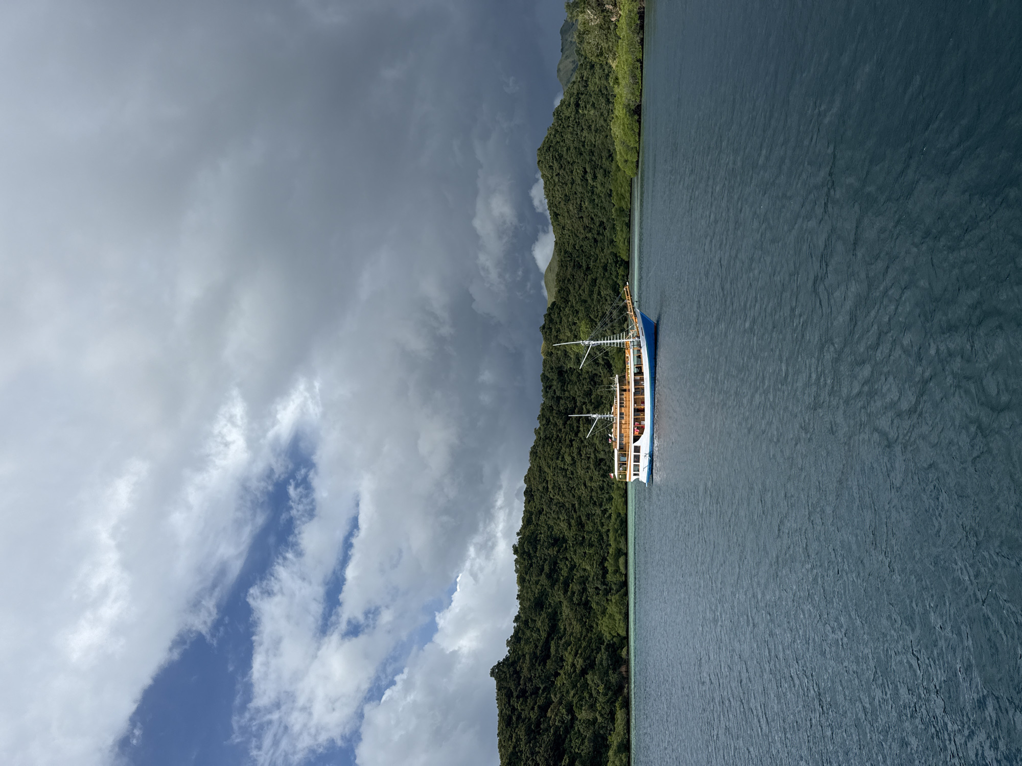 The image depicts a boat sailing on the water near Rinca Village, Indonesia. The sky is filled with clouds. The land is covered with dense green vegetation. The water appears calm.