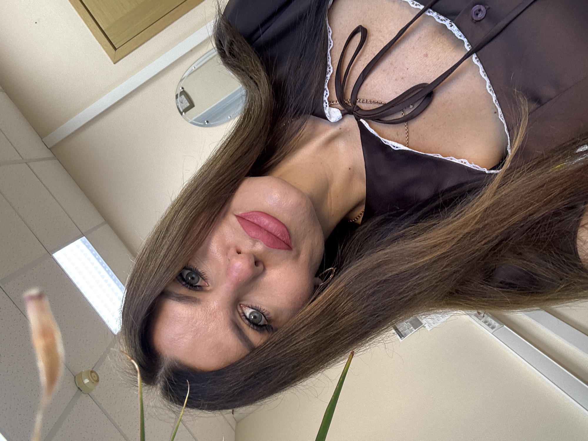In a ceiling-lit room, a woman with long brown hair, wearing a dark brown outfit with white lace trim and a gold chain necklace, poses for a selfie. She has light-colored eyes and is wearing pink lipstick. The background shows a beige wall, a white baseboard, and a wooden door. A plant stem is visible in the upper left corner of the image. The setting is likely an indoor office or similar space.