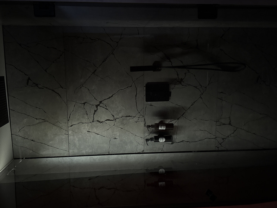 The image depicts a modern, dimly lit shower enclosure within an interior setting in Kyiv, Ukraine. The walls are clad with large, grey, marble-patterned tiles featuring prominent dark veining. A clear glass shower screen separates the shower area, reflecting interior elements including two pump dispenser bottles mounted on the tiled wall. The bottles are one dark, cylindrical unit and one amber-brown, both with white labels. To their right, a black square-shaped control panel is affixed to the wall. Further right, a black handheld showerhead is suspended from its hose. The scene is devoid of individuals or active interactions. The environment suggests a contemporary bathroom design with low ambient lighting creating shadows and reflections on the polished surfaces. No exterior visual cues indicate a specific location within Kyiv.