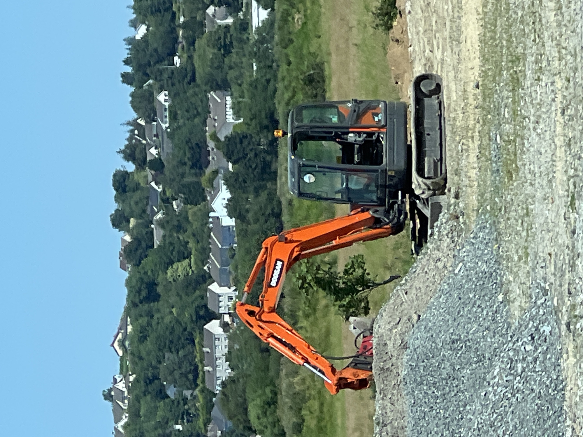 A bright orange excavator, branded with "DOOSAN", sits in a construction area in Letterkenny, Ireland. The excavator is positioned on a dirt and gravel surface, with a large pile of grey gravel to its left. The machine's arm is raised, and it appears to be equipped with a breaker attachment. In the background, a green, treed hillside is dotted with numerous white-roofed houses and buildings, suggesting a suburban or rural edge of the city. The sky is a clear, bright blue. The scene suggests ongoing construction work in a residential area.
