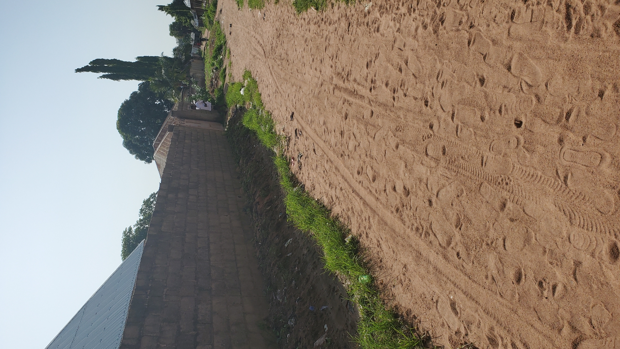 The image depicts a dirt road in the city of Jalingo, Nigeria. A tall, grey brick wall runs along the left side of the road, bordering what appears to be a residential or commercial area. Beyond the wall, a corrugated metal roof of a building is visible. The road itself is sandy and covered with numerous footprints and tire tracks, suggesting it is a frequently used pathway. To the right of the road, sparse vegetation and some scattered debris can be observed. In the distance, several trees, including a tall, slender one with multiple spires, are visible against a clear sky. The overall scene suggests a dry, dusty environment typical of certain areas in Jalingo. There are two individuals visible in the distance, one standing near the wall and another walking further down the road. Their presence indicates activity and life within the scene.