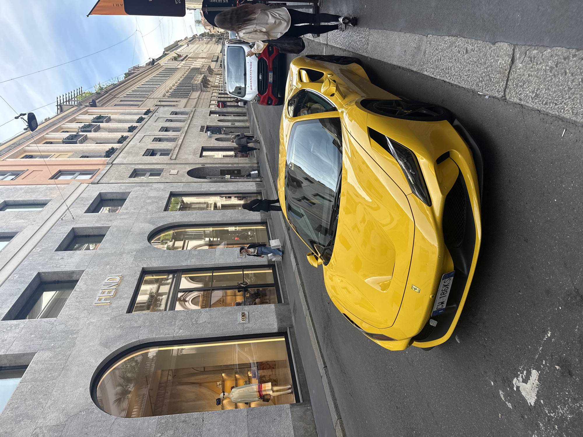 The image captures a street scene in Milan, Italy. A bright yellow Ferrari is parked along the right side of the street. Next to the car is a white van with red accents. A person is standing near the van. Pedestrians are walking on the sidewalk, including one in front of the car. The building on the left side of the street houses a FENDI store. Buildings rise on the opposite side of the street. The sky is visible in the upper portion of the frame.