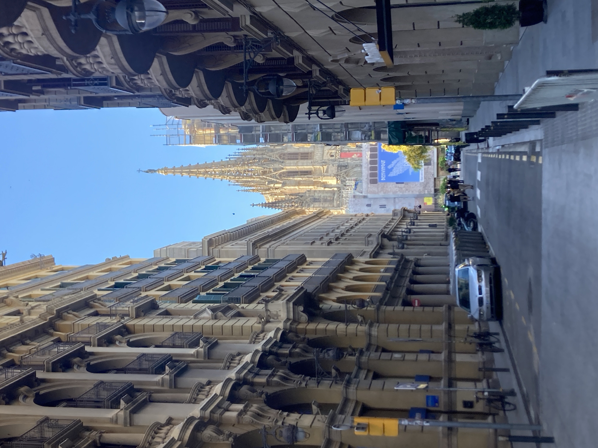 The image shows a street scene in Barcelona, Spain, looking towards the Barcelona Cathedral. The street is lined with tall, ornate buildings with arched windows and balconies. A silver car is parked on the side of the street, and several bicycles are also visible. In the background, the spires of the Barcelona Cathedral dominate the skyline. The sky is clear and blue. The overall impression is one of a historic European city on a sunny day. The scene is typical of the Gothic Quarter of Barcelona.