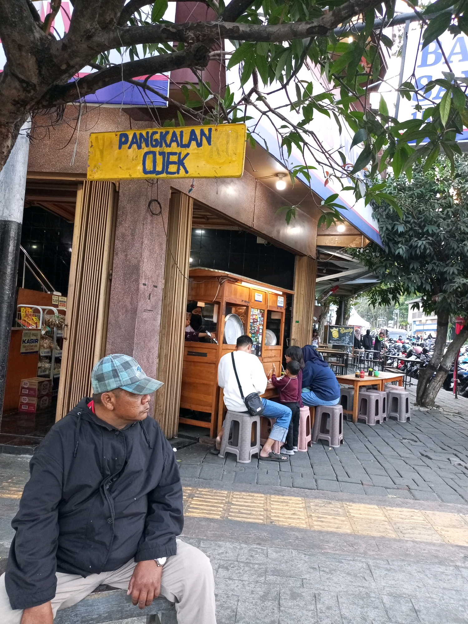 A man wearing a plaid cap and black jacket sits on a bench in front of a building with a yellow sign that reads "Pangkalanj Ojek." The sign is in blue lettering. The building appears to be a food stall or restaurant. Three people are seated at a table in front of the building, and others are visible in the background. A street with parked motorcycles is also visible. The scene is outdoors, on a paved area next to the building. The overall impression is of a typical street scene in a city.
