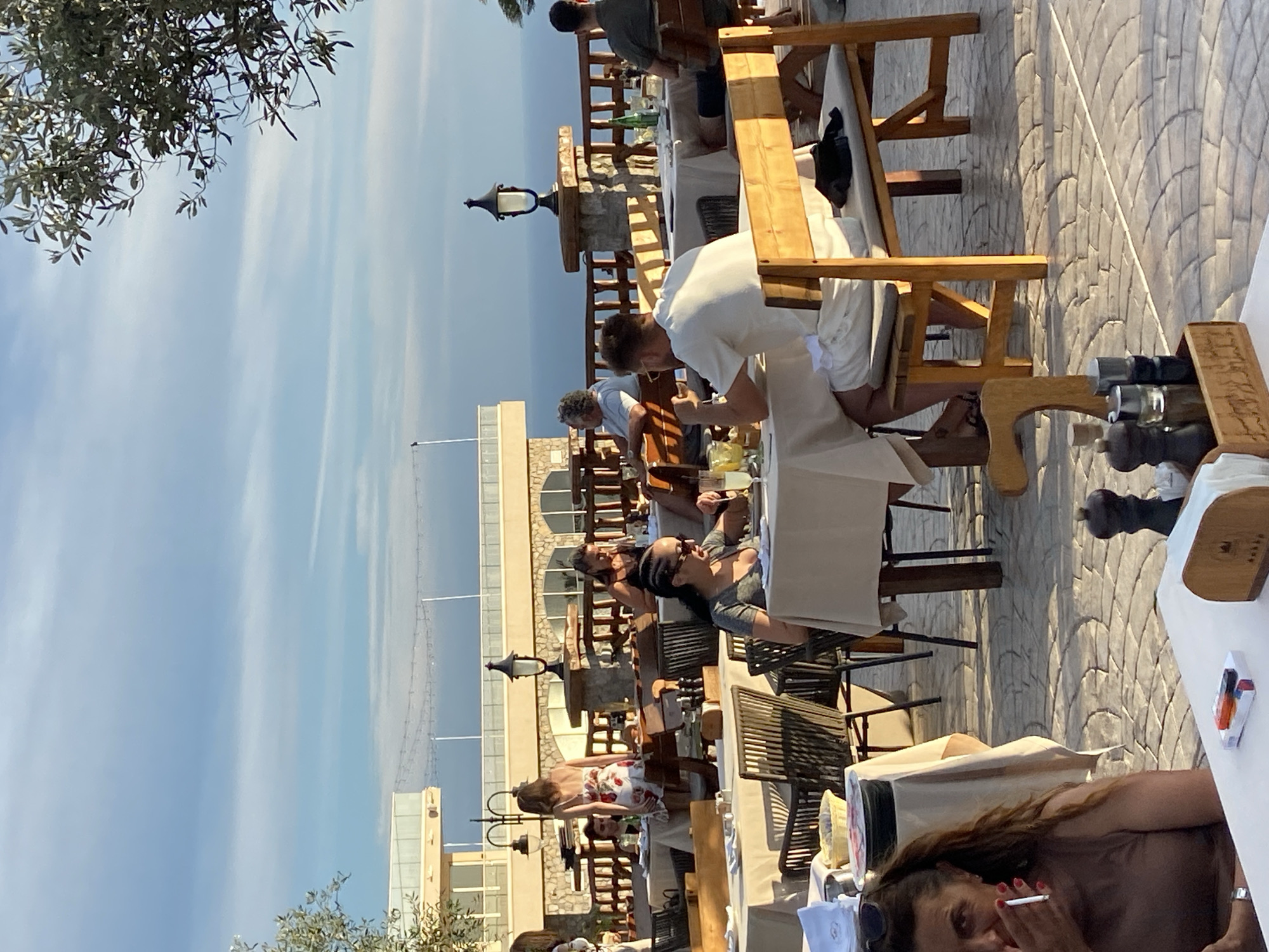 The image depicts an outdoor dining area of a restaurant, possibly located in Kovačko Polje, Montenegro, overlooking the sea under a clear sky with scattered clouds. Several tables are set with white tablecloths and chairs. People are seated at various tables, engaged in dining and conversation. In the foreground, a woman with dark hair is smoking a cigarette, holding it to her mouth. She is wearing a brown tank top. To her right, a wooden salt and pepper grinder set is visible on a white tablecloth. Further back, a man in a white t-shirt and shorts sits at a table with a wooden bench. Another table features a woman with braided hair and sunglasses, eating. Behind them, other patrons are seen at their tables. The restaurant structure, with its stone facade and arched windows, is visible in the background, along with a railing and decorative lighting. The ground is paved with stone tiles.