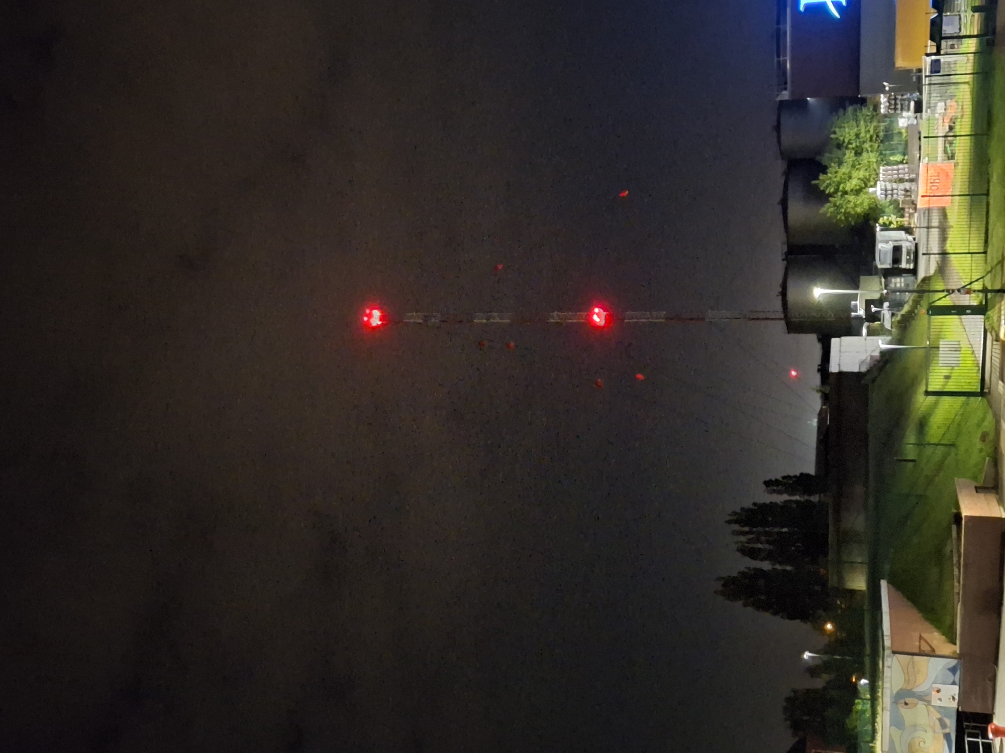 Here's a description of the image:
The image shows a night scene, likely in an urban or industrial area. In the foreground, a tall radio tower stretches vertically into the night sky. The tower is marked with several red lights. To the right, there is a building with a neon blue sign. Adjacent to the building, there are industrial-looking tanks. A chain-link fence separates the tanks from a grassy area. On the other side of the tower, the night sky is dark, possibly due to heavy cloud cover or smog. The image is taken from a fixed position, likely at ground level.