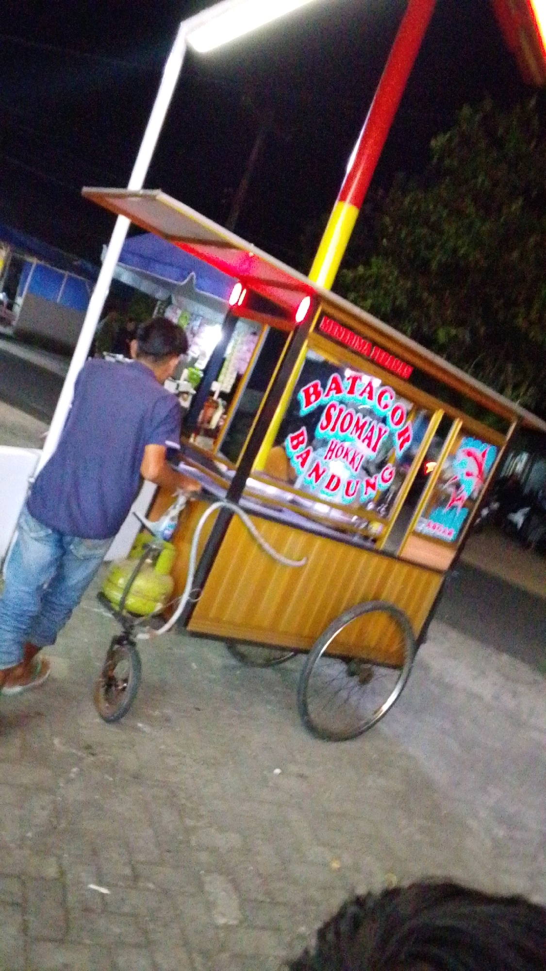 The image depicts a street food cart with "BATAGOR SIOMAY HOKKI BANDUNG" written on it. The cart is yellow and has red accents on top. A person wearing a blue t-shirt and jeans is standing near the cart, seemingly operating it. There is a green gas cylinder attached to the cart. The ground is paved with bricks. The scene appears to be at night, indicated by the dark background and the lighting from the cart and a nearby lamp post. The location is identified as Desa Karangbenda, Indonesia.