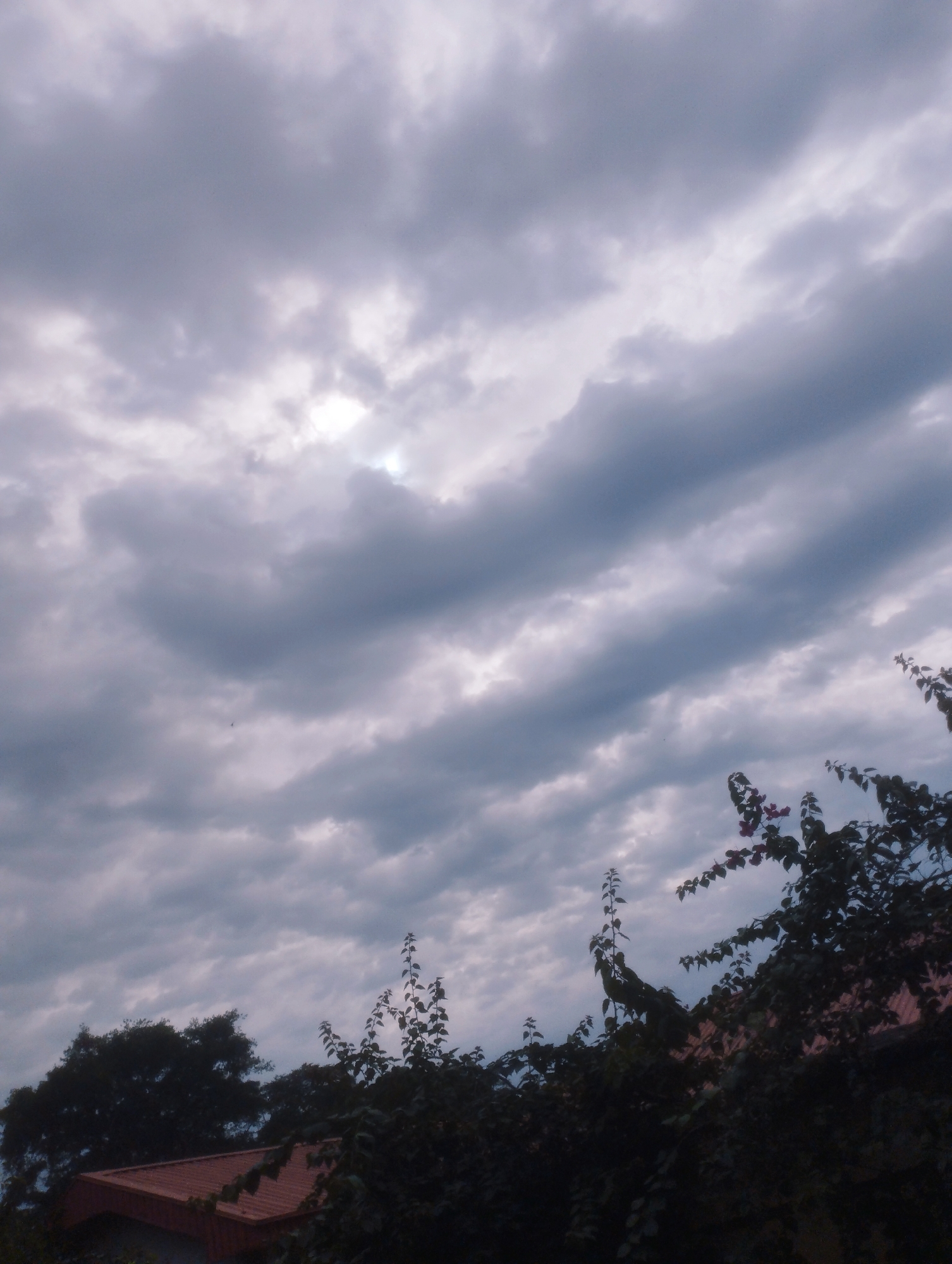 A low-angle, outdoor shot captures a dramatic, cloudy sky over what appears to be a residential area. The clouds are thick and layered, with streaks of lighter gray and white suggesting an overcast or stormy condition. A patch of brighter light, possibly the sun, peeks through a gap in the clouds near the top center. In the foreground, dark silhouettes of trees and foliage obscure the lower portion of the image, with hints of a reddish-brown roofline visible beneath. The scene evokes a somber or dramatic mood, typical of pre-storm weather. It is not possible to determine the specific location within Jos, Nigeria from the visual information provided.