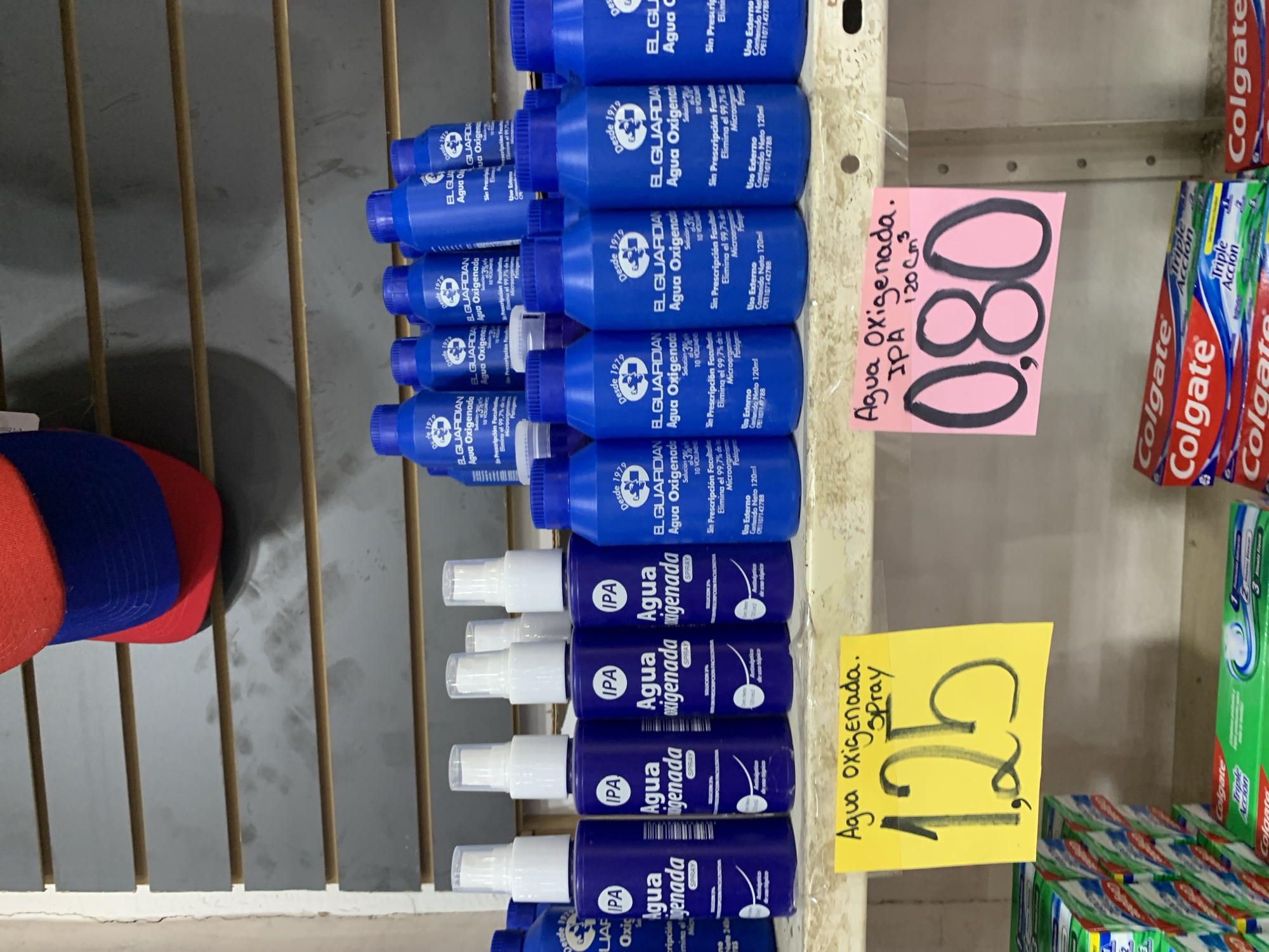 The image shows a shelf in a store, likely a pharmacy or small supermarket, with various products displayed. In the foreground, on the left, a yellow price tag reads "Agua oxigenada. Spray" with the price "1,25" handwritten in large black numbers. To the right of this, a pink price tag reads "Agua Oxigenada. IPA 120 Cm³" with the price "0,80" in large black numbers. Below the yellow tag are stacks of Colgate toothpaste boxes. Below the pink tag are more stacks of Colgate toothpaste boxes, some with red and white packaging and others with green and white packaging.
On the shelf above, there are rows of blue plastic bottles with white spray tops. These bottles are labeled "IPA Agua Oxigenada". Behind these, in the upper part of the image, are more blue bottles, some with white caps and others with blue caps, also appearing to be some form of antiseptic or cleaning product, with labels that include "EL GUARDIAN Agua Oxigenada".
At the very top of the image, a red, white, and blue baseball cap is visible, resting on a shelf or display. The overall environment appears to be an indoor retail setting with utilitarian shelving. The lighting is somewhat harsh, suggesting artificial store lighting. The location is identified as Santa Ana, Venezuela.
