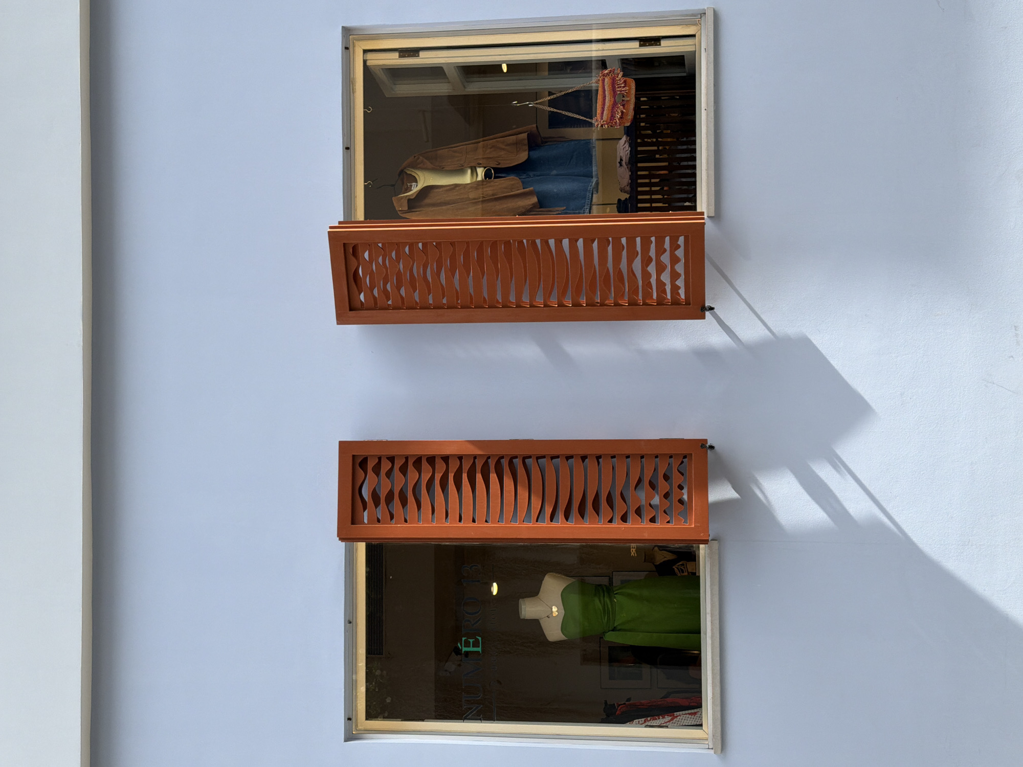 Two windows of a shop, one above the other, face the viewer. Both windows are mostly visible with an opening at the top, both of which have decorative brown panels on top that extend like awnings. The wall is a pale blue color. The top window features a mannequin dressed in a brown jacket, blue jeans and a tan shirt. A purse also hangs by the upper left of the window. The lower window has a mannequin in a green dress. A logo visible on the window reads “NUMERO 13”.