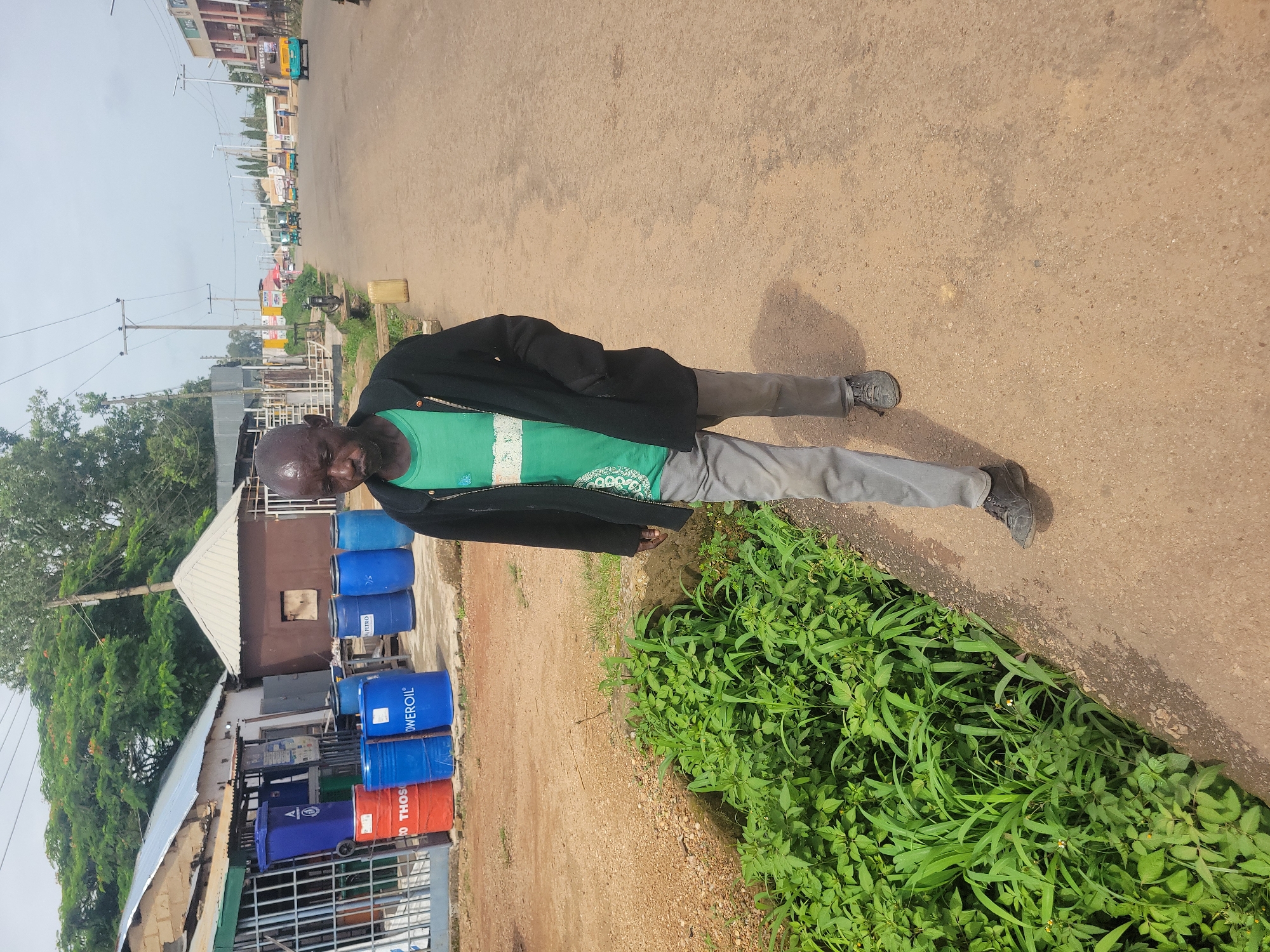A bald, dark-skinned man walks on a dirt road in Jos, Nigeria. He is wearing a green t-shirt with a white stripe and a dark jacket. Gray pants and dark shoes complete his attire. The man is walking away from the camera, with his head slightly bowed. The road is lined with lush green vegetation on the left side and a few buildings on the right. The sky is overcast. In the background, there are several buildings, some with corrugated iron roofs, and a few vehicles, including a yellow and blue tuk-tuk. Several blue and red barrels are visible in front of one of the buildings. The overall impression is one of a typical street scene in a Nigerian city.
