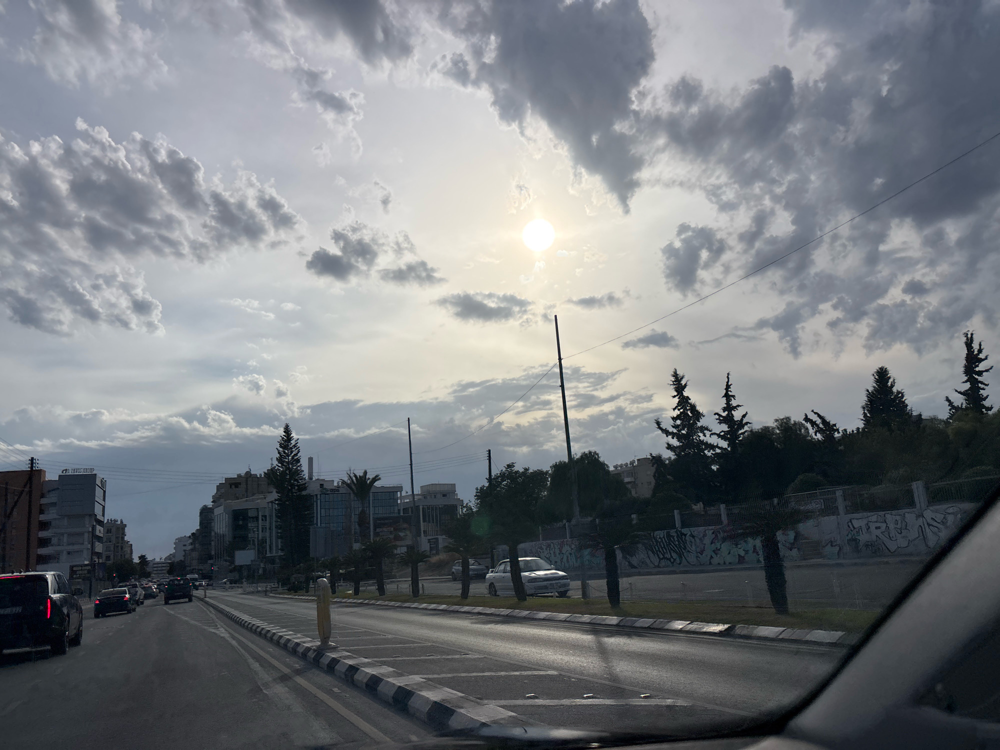 The image captures a view from a vehicle traveling on a multi-lane road in Agios Athanasios Municipality, Cyprus. The sky is filled with dramatic, partly cloudy formations, with the sun visible as a bright, diffused orb through the haze. On either side of the road are modern buildings and trees, including palm trees and coniferous trees. The road is lined with pedestrian sidewalks and traffic markings. Several vehicles are visible on the road, indicating moderate traffic. Graffiti can be observed on a wall in the background. The overall impression is that of a city street scene under an overcast sky.