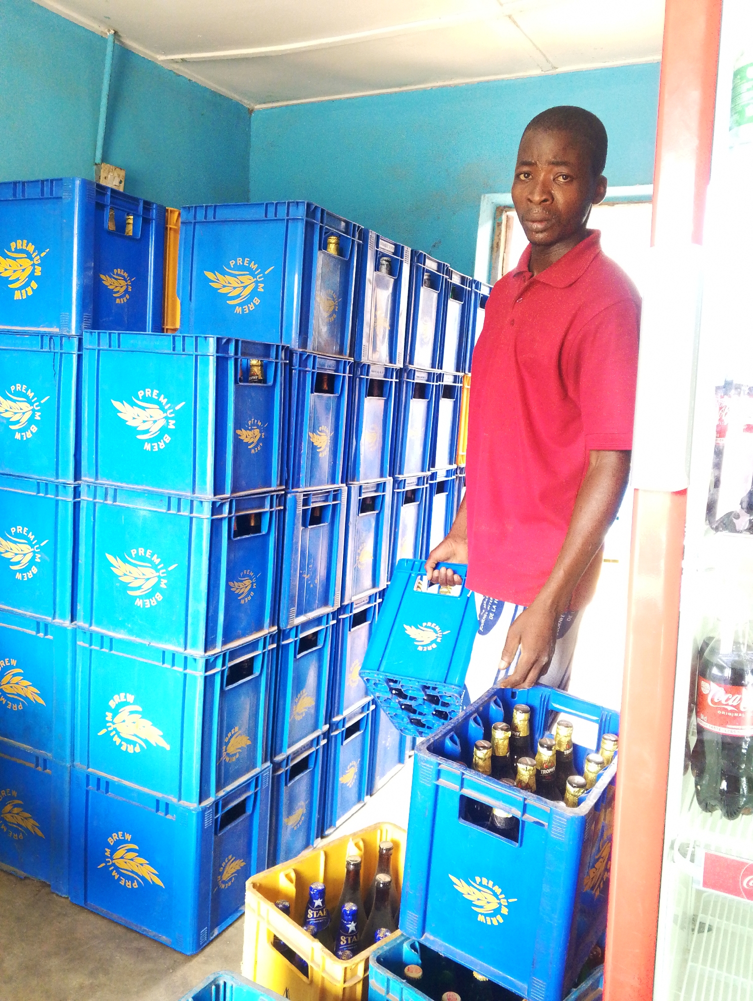 A man stands in a small, brightly lit room, likely a store or storage area, surrounded by stacks of blue and yellow plastic crates filled with bottled beverages. The man is wearing a red polo shirt and light-colored shorts or trousers. He is holding a blue crate filled with bottled beverages, which appear to be beer, with gold caps. The crates are marked with "PREMIUM BREW" and a logo of wheat or barley stalks. In the background, a stack of blue crates dominates the left side of the frame. A yellow crate can be seen at the base of the stack. To the right, a refrigerator is visible with Coca-Cola bottles inside. The room is painted blue, with a white ceiling.