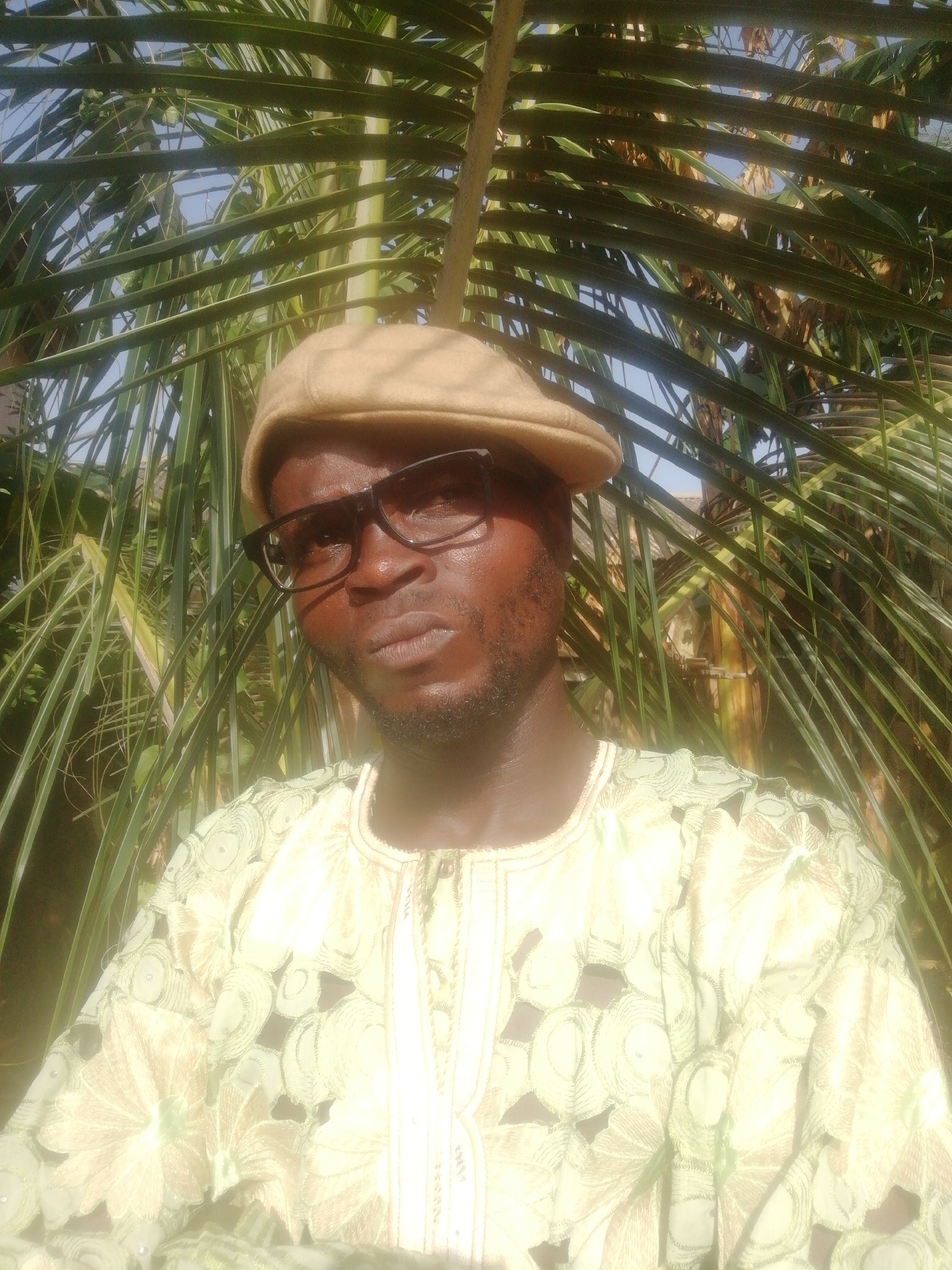 A close-up, brightly lit outdoor portrait features a man against a backdrop of lush green palm fronds. The man, with dark skin, is centered in the frame, looking directly towards the viewer. He wears a light brown or tan flat cap and dark-framed glasses. His lips are slightly pursed, giving him a neutral to slightly serious expression. He has a short beard.
He is dressed in a light-colored, possibly cream or light green, traditional garment with intricate cut-out patterns that appear floral or lace-like, possibly embroidery, over a darker fabric underneath. The patterns are prominent on the shoulders and chest area.
The background is dominated by numerous overlapping green palm leaves, suggesting a tropical or garden setting. Patches of bright blue sky are visible through gaps in the foliage, indicating clear weather and a sunny daytime setting. The strong sunlight creates highlights on the man's skin, glasses, and hat, and illuminates the green leaves. There is no visible text in the image.