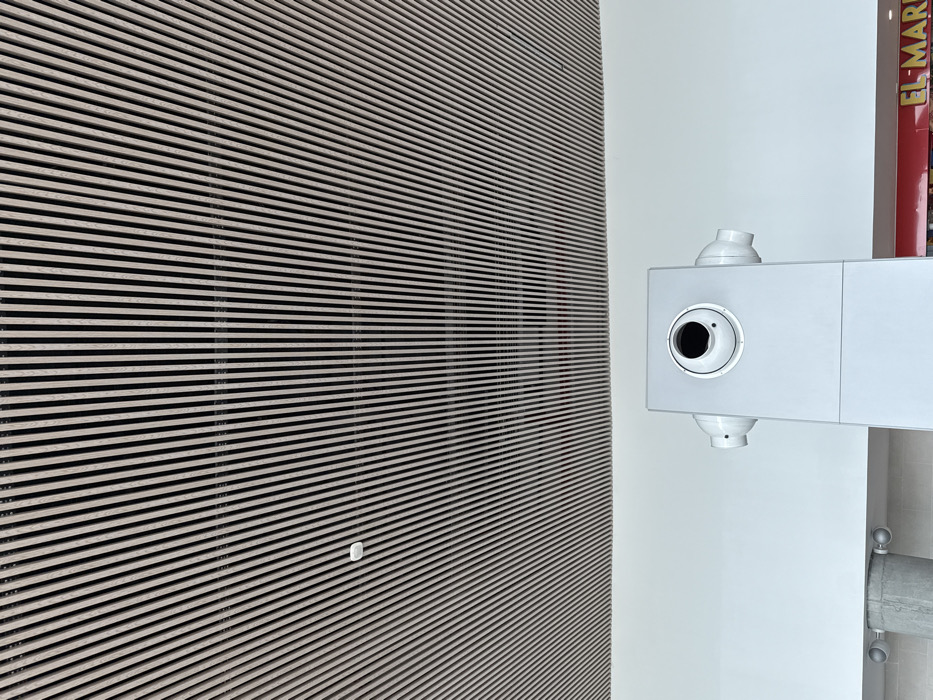 The scene is located in the city of AMCO, Area Metropolitana Centro Occidente, Colombia.
The image presents an indoor architectural perspective, dominated by a diagonally slatted ceiling composed of light-toned rectangular elements. A small, white, circular sensor or detector is affixed to this ceiling in the upper-left quadrant. Below the ceiling, a clean white wall occupies the middle section of the frame. A prominent light-gray, rectangular architectural element or column stands in the lower-center, extending upwards from the bottom edge. This element features a large circular recess with a dark, possibly empty, interior, and a smaller circular component offset to its right, suggestive of a camera or sensor housing. Two symmetrical, light-gray cylindrical fixtures protrude horizontally from either side of this rectangular element. To the left of this central element, a cylindrical concrete column is partially visible, featuring two circular lights or fixtures. In the lower-right corner, a section of a red sign with white capital letters, partially reading "EL MAR," is visible, indicating a commercial establishment. No individuals are present in the frame.