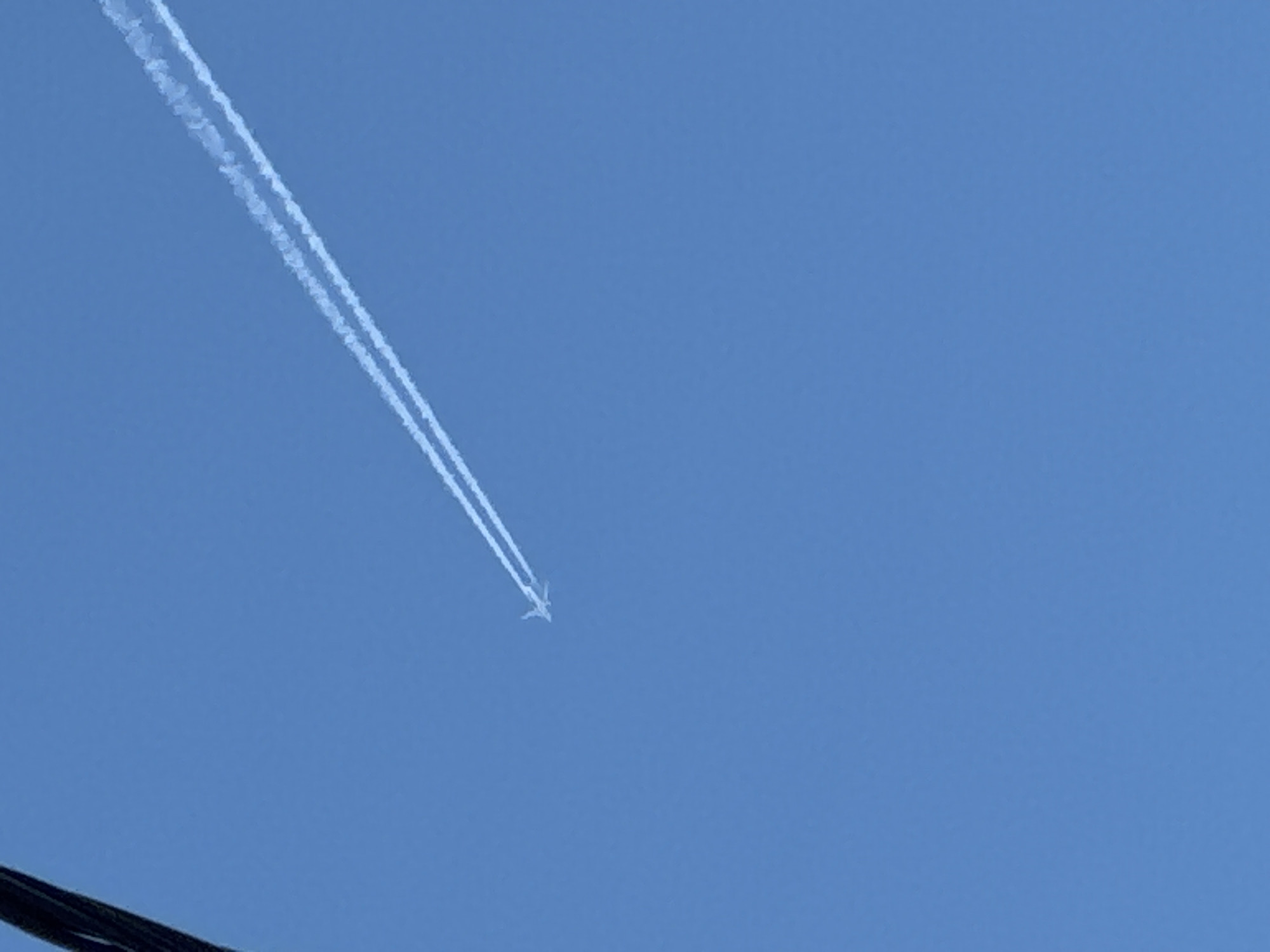 A contrail is visible in a clear blue sky. The contrail is made up of two parallel lines extending from the top left of the frame downwards towards the center where a small plane is visible. A black wire is at the bottom left. The location of this image is indeterminate.