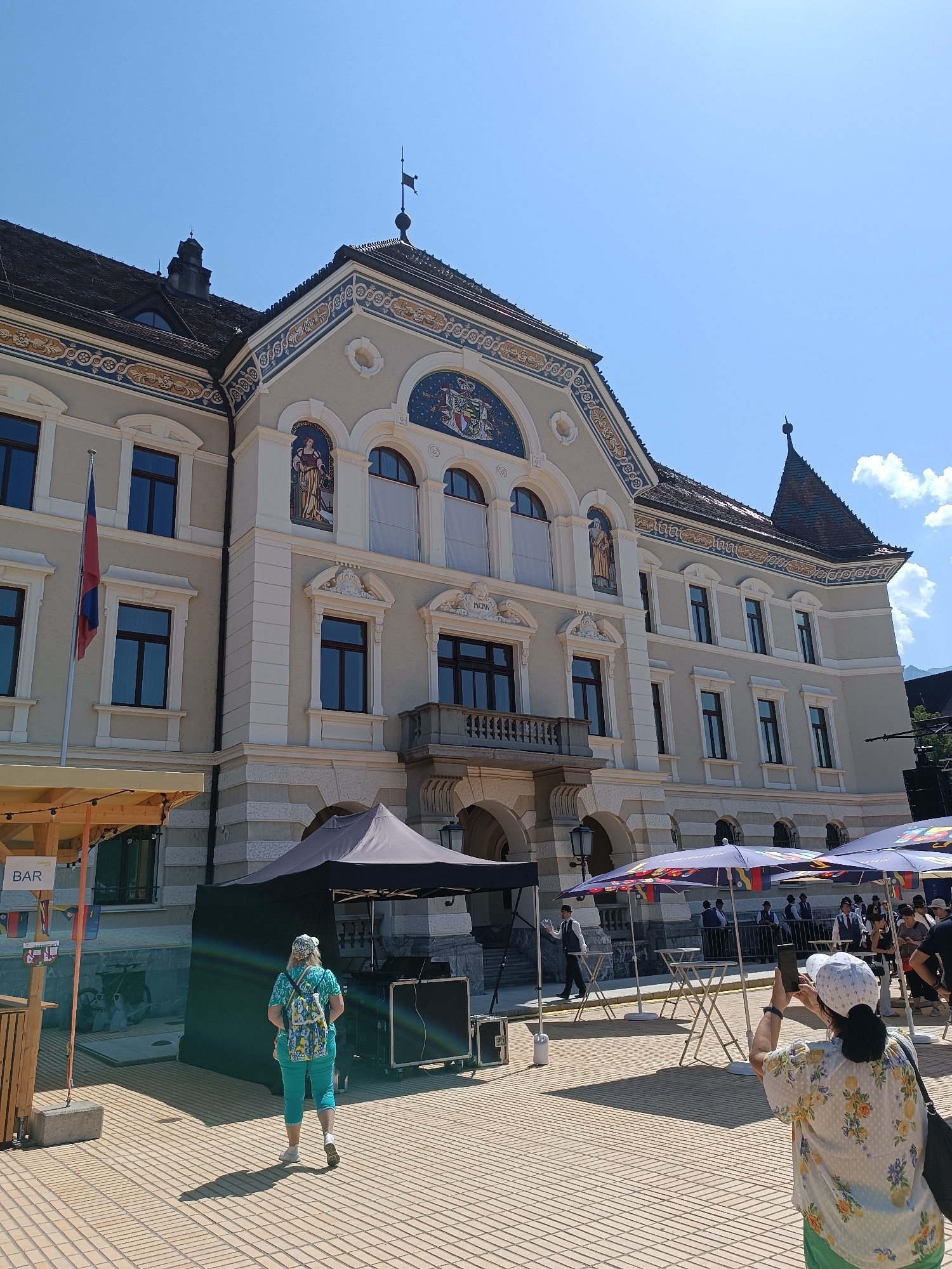 The image depicts the exterior of the Vaduz Town Hall in Vaduz, Liechtenstein, on a sunny day. The building features intricate architectural details, including decorative friezes, arched windows, and a tiled roof. In front of the building, there is an outdoor event setup. Several tables and chairs are arranged, some covered by colorful umbrellas. A black tent is set up, possibly for an event or vendor.

In the foreground, a woman wearing a patterned cap, a floral print shirt, and green pants is taking a picture with her smartphone. She is facing away from the camera, towards the building. To her left, another person wearing a backpack and teal-colored leggings is walking away from the camera.

Further in the background, several individuals are visible, some in formal attire, possibly part of an event staff or attendees. A flag is planted near the building. The ground is paved with interlocking bricks. The sky is clear blue with a few white clouds visible on the right side of the frame. A "BAR" sign is visible on a wooden structure to the left.