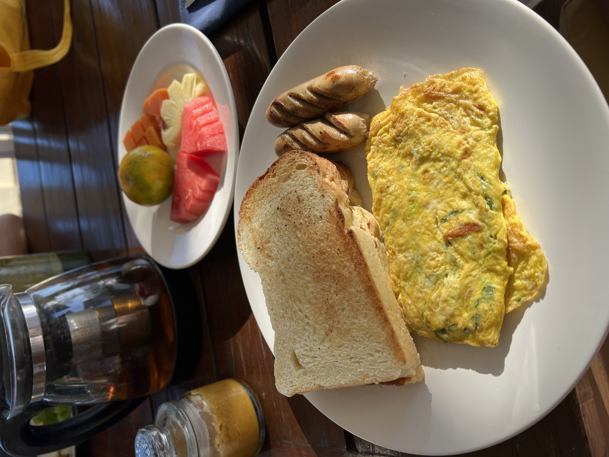 A breakfast setting is depicted on a wooden table in Busungbiu, Indonesia. In the foreground, a white plate holds a folded omelet with visible green vegetable pieces, two grilled sausages, and two slices of toasted white bread. To the left, a small glass jar contains a golden condiment, likely jam or honey, and a clear glass teapot filled with amber-colored tea is positioned behind it. In the background, another white plate features a colorful fruit salad including watermelon slices, orange segments, diced papaya, and pineapple. A bright yellow cloth bag rests further back, and a blue napkin is visible on the right side of the frame. The scene is illuminated by natural sunlight, casting distinct shadows on the table.