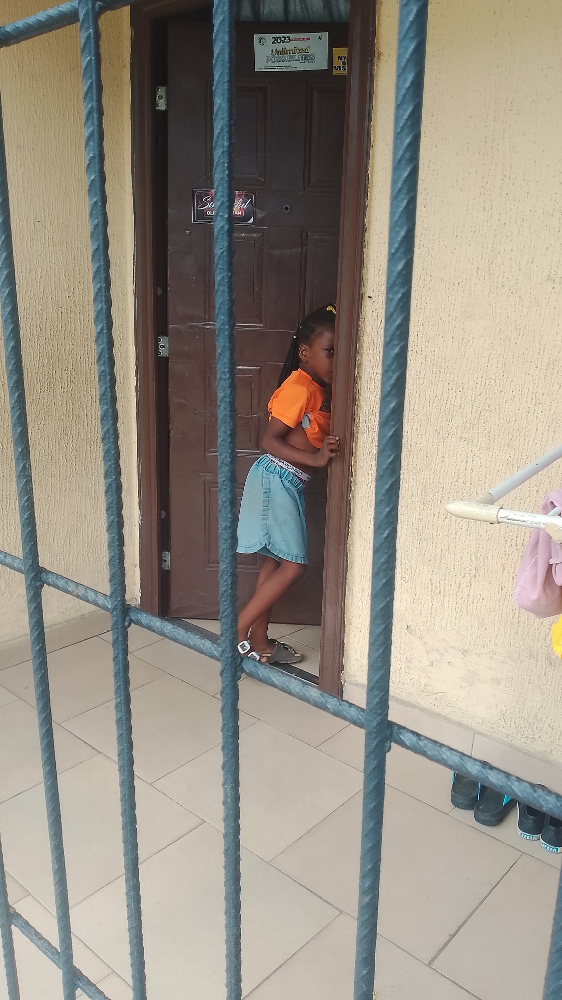 The image shows a young Black girl standing at a doorway, peeking around the edge of the doorframe into what appears to be a room. She is wearing an orange t-shirt that is pulled up, revealing her midriff, and a light blue denim skirt. She has her hair in braids with yellow ties. She is barefoot, wearing sandals. The doorway is dark brown and has some stickers on it. One sticker reads "2023 Unlimited POSSIBILITIES." Another sticker has "Ste ful" written on it. The girl's stance and the way she is peering suggest curiosity or a playful moment. The setting appears to be outdoors, possibly a porch or patio, with a tiled floor and a textured wall. In the background, a clothesline with some pink and yellow fabric is visible. Several pairs of black sandals are lined up on the floor near the wall. The lighting suggests it is daytime.