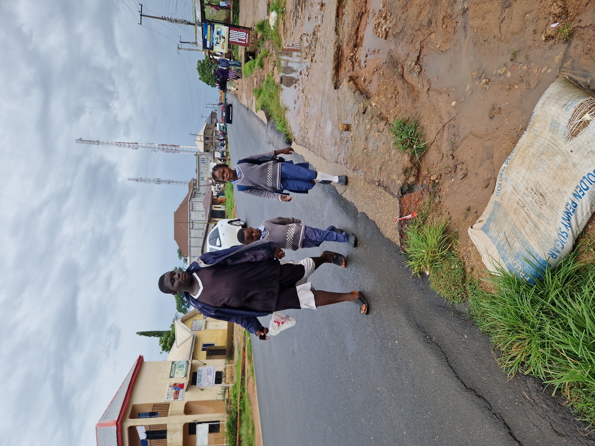 On Rukuba Road, Nigeria, a person walks alongside two children on a wet asphalt road. The adult, wearing a dark shirt and blue jacket over shorts, carries a white plastic bag. The two children, dressed in school uniforms consisting of grey sweaters, white shirts, blue skirts for the girl, and blue trousers for the boy, walk together, with the boy holding the adult's hand. The girl carries a blue backpack. The environment is characterized by a cloudy sky and a damaged road with puddles and exposed earth. Buildings and telecommunications towers are visible in the background. A large, damaged sack, possibly containing sugar, lies on the side of the road near overgrown grass. The scene suggests a typical day on a Nigerian road, possibly after rain, with a family walking home.