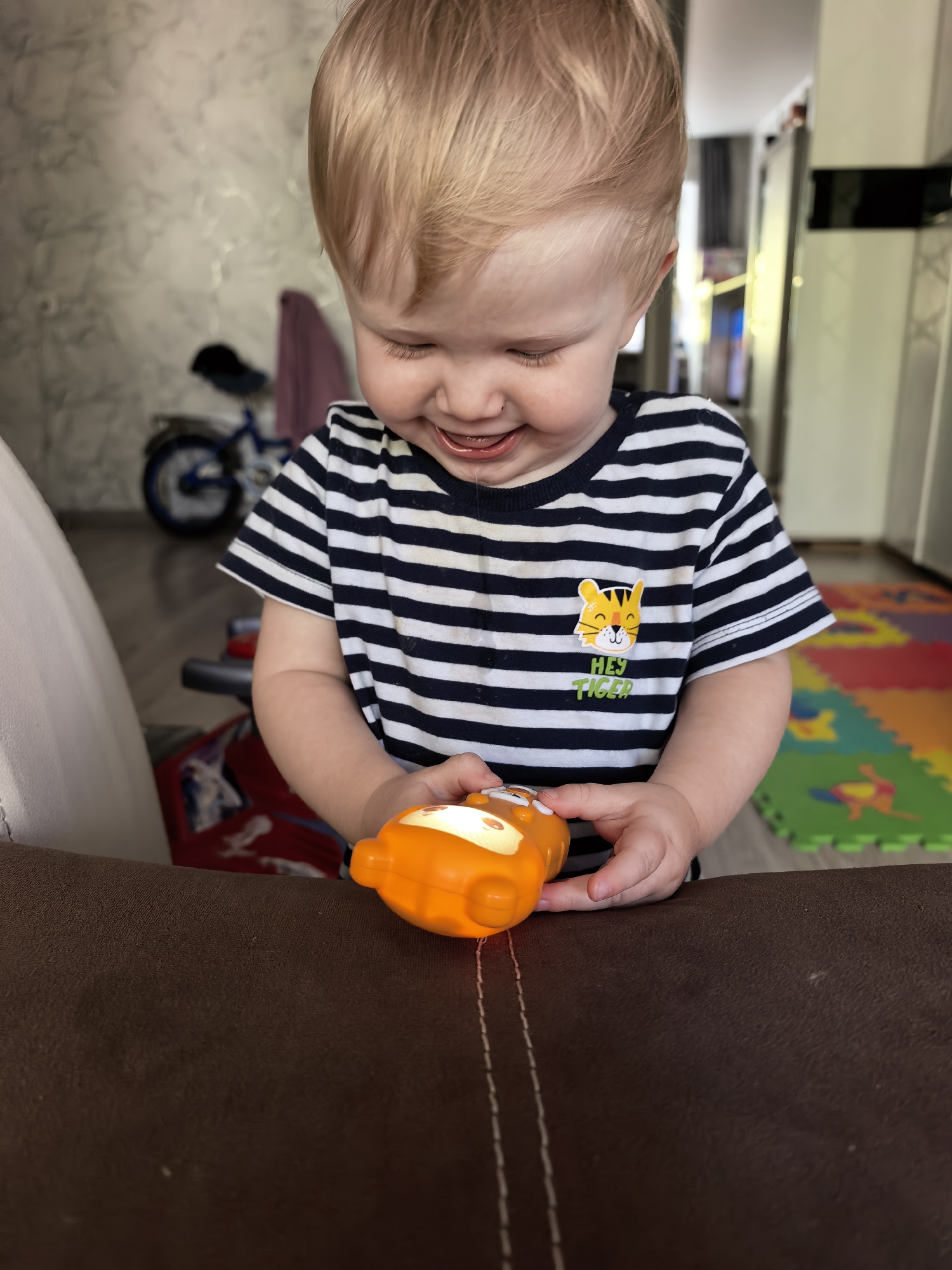 A young child with blonde hair and a striped shirt is playing with an orange toy. The child is looking down at the toy with a smile. In the background, there is a bicycle, a play mat with colorful shapes, and kitchen appliances. The image appears to be taken indoors, possibly in a home, in the city of Sterlitamak, Russia.