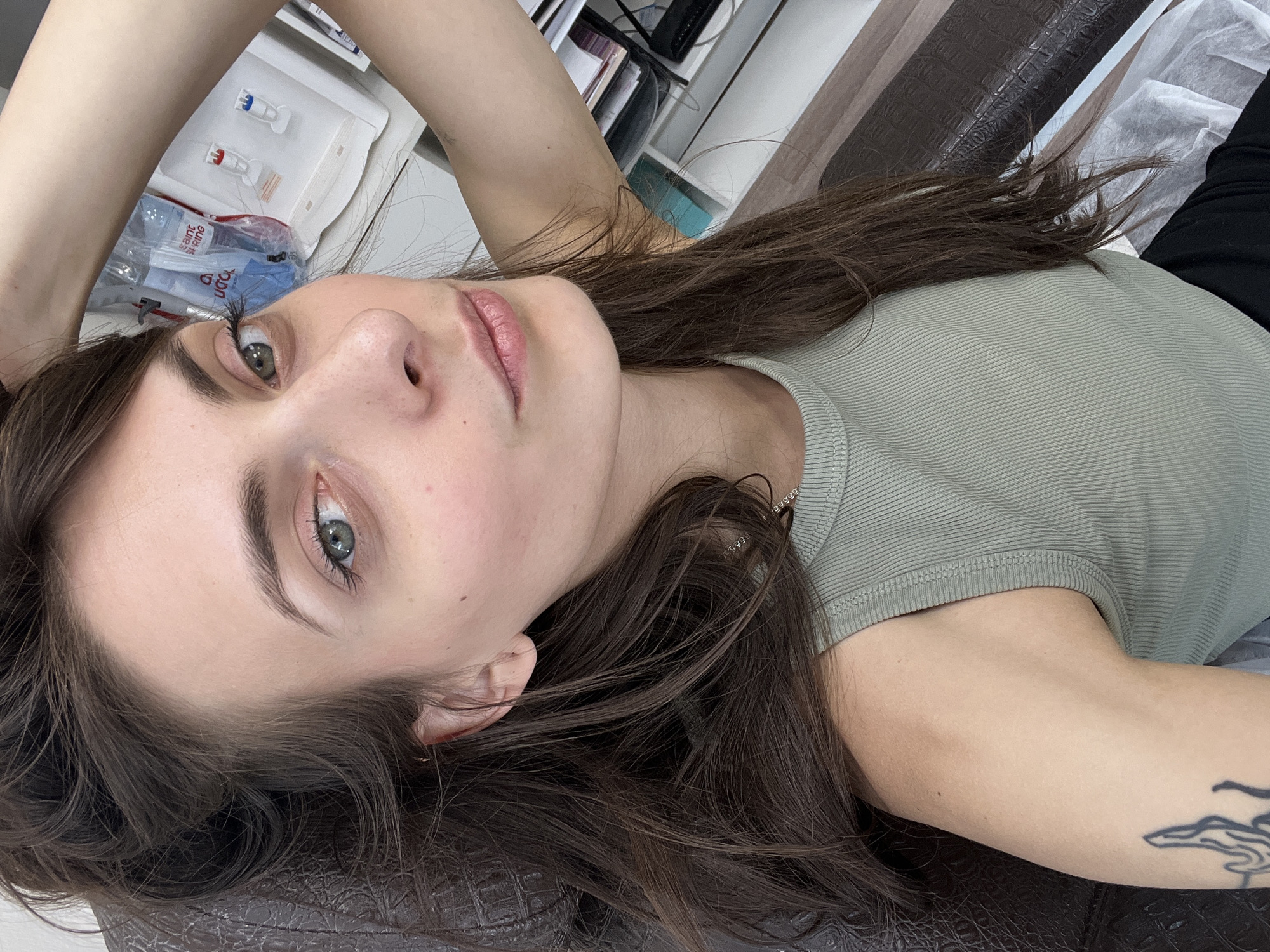 A young woman with long, dark brown hair and light green or hazel eyes is captured in a close-up, horizontal shot. She is lying on her back, looking directly at the camera with a neutral, serene expression. She wears a light olive green ribbed tank top or camisole, and a black tattoo with flowing, abstract designs is visible on her left upper arm. Her left arm is raised above her head, while her right arm is out of frame.
She is resting on a dark brown, textured surface that resembles patterned leather, possibly an examination table or salon chair. The background indicates an indoor setting, likely a beauty salon, aesthetic clinic, or a similar treatment room in Krasnoyarsk, Russia. Various medical or beauty supplies are visible on a white surface to her left. These include small plastic vials with distinct red and blue caps. A plastic bag containing other items also shows partial text, including "saint," "STOPPING," and "PUDRA" (Russian for powder), reinforcing the context of an aesthetic or cosmetic procedure. The room is well-lit, suggesting either natural daylight or ample artificial lighting.