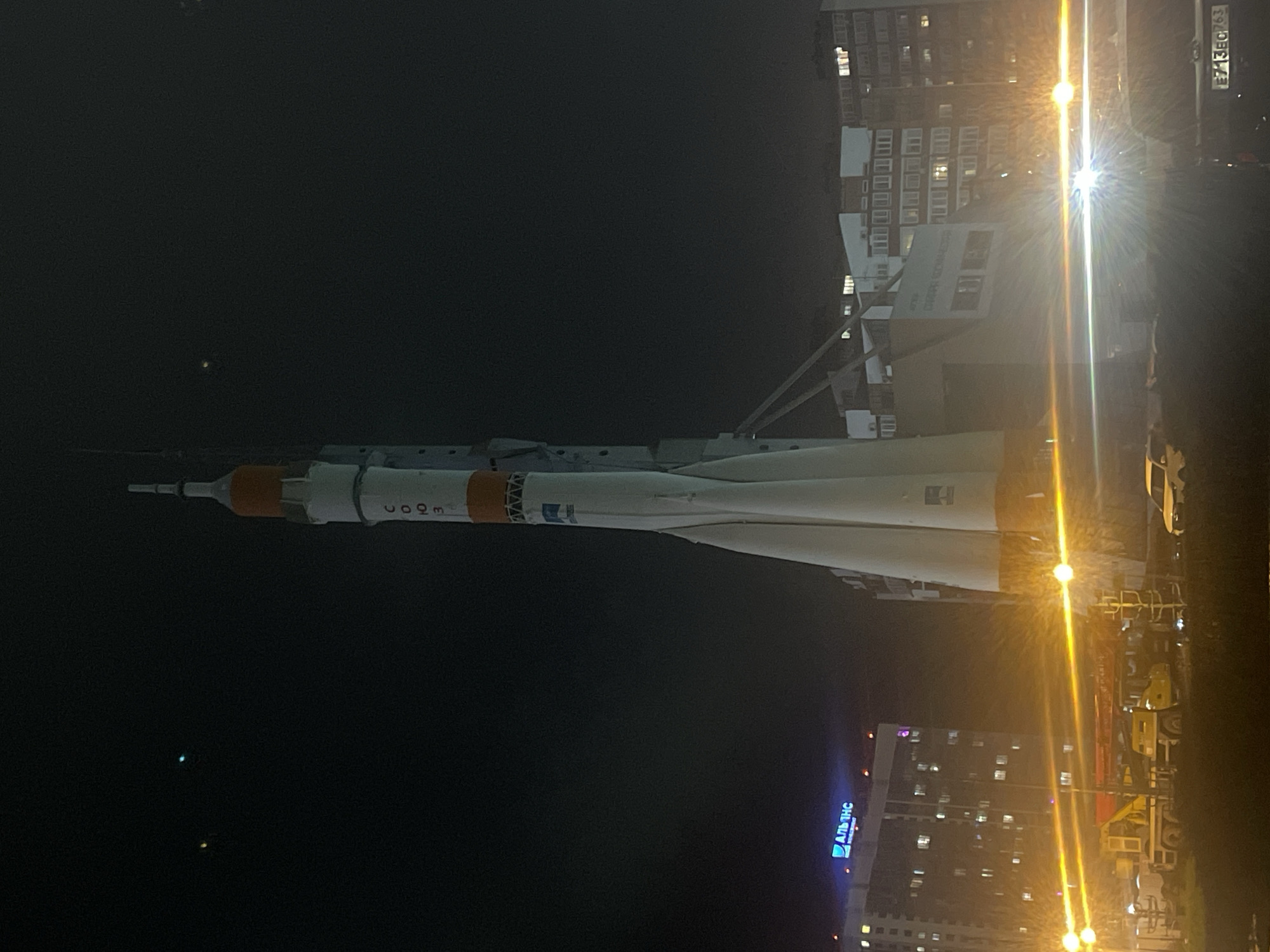 The image shows a Soyuz rocket monument on display at night in Novokuybyshevsk, Russia. The rocket is illuminated by streetlights, and nearby buildings are visible. A vehicle is parked on the side of the road. The sky is dark.