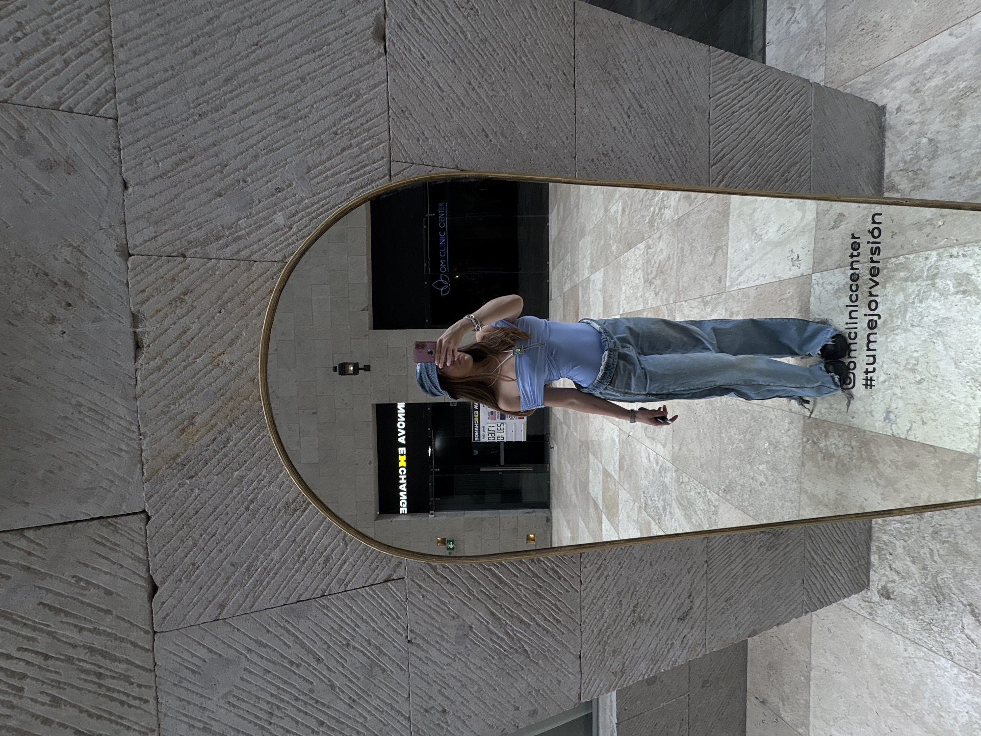A full-length mirror in an arched gold frame stands outside a building in Ciudad López Mateos, Mexico. The mirror reflects a young woman standing and taking a selfie with a phone, partially obscuring her face. She is wearing a light blue top, jeans, and a blue hat. The reflection also shows the entrance to a business called "OM Clinic Center," and a currency exchange with reversed signage. The surrounding architecture consists of textured stone walls and tiled flooring. In the lower right corner of the mirror, the words "@omcliniccenter #tumejorversión" are displayed.