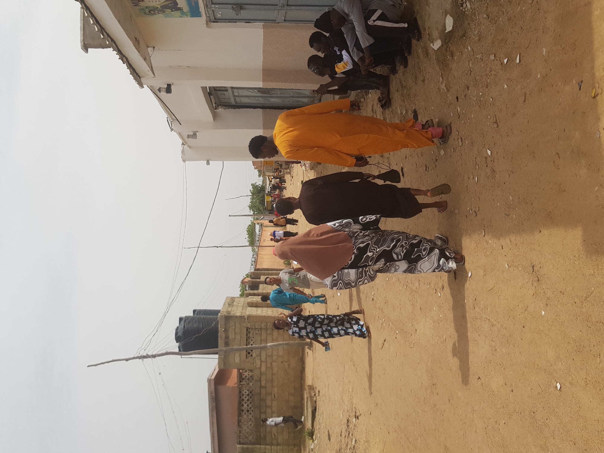 A wide shot shows a street scene in Dabawa, Nigeria. The street is unpaved and made of packed earth. Several people are walking on the street. From right to left, the people are clustered into groups. One group includes at least six men near a building who are crouching. Another man is wearing an orange garment. Another man is wearing a dark brown robe. A woman is wearing a brown hijab and a black and white patterned dress. Further down the street, more people can be seen. Buildings line the left of the street, and one of them has black water tanks. Power lines are strung between poles. The sky is hazy.